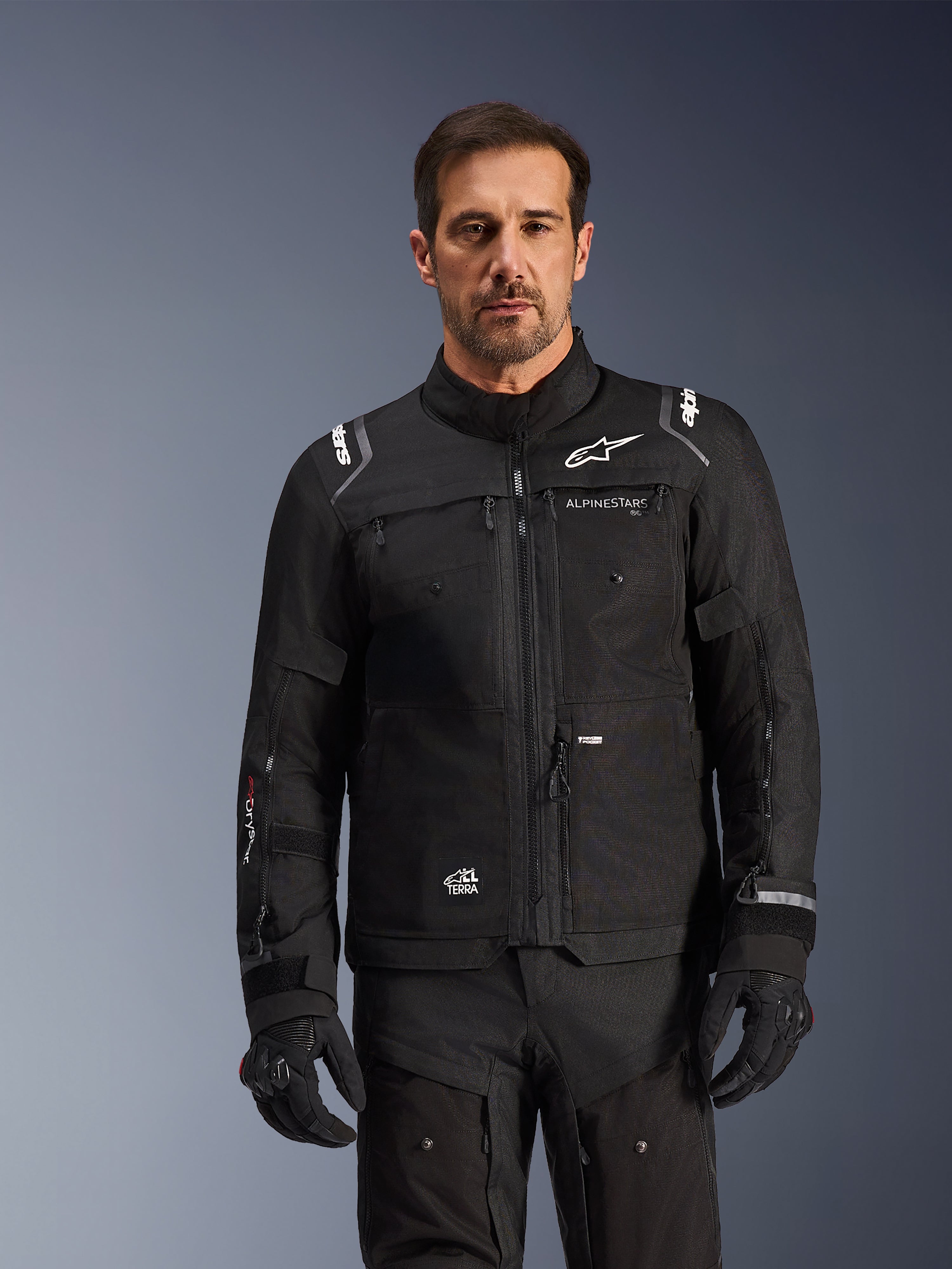 Alpinestars Cusco Drystar®-jacka, svart motorcykeljacka, bärs av en modell med matchande svarta handskar och byxor, med teknisk textilkonstruktion och vita Alpinestars-logotyper, designad för äventyrstouring på landsväg