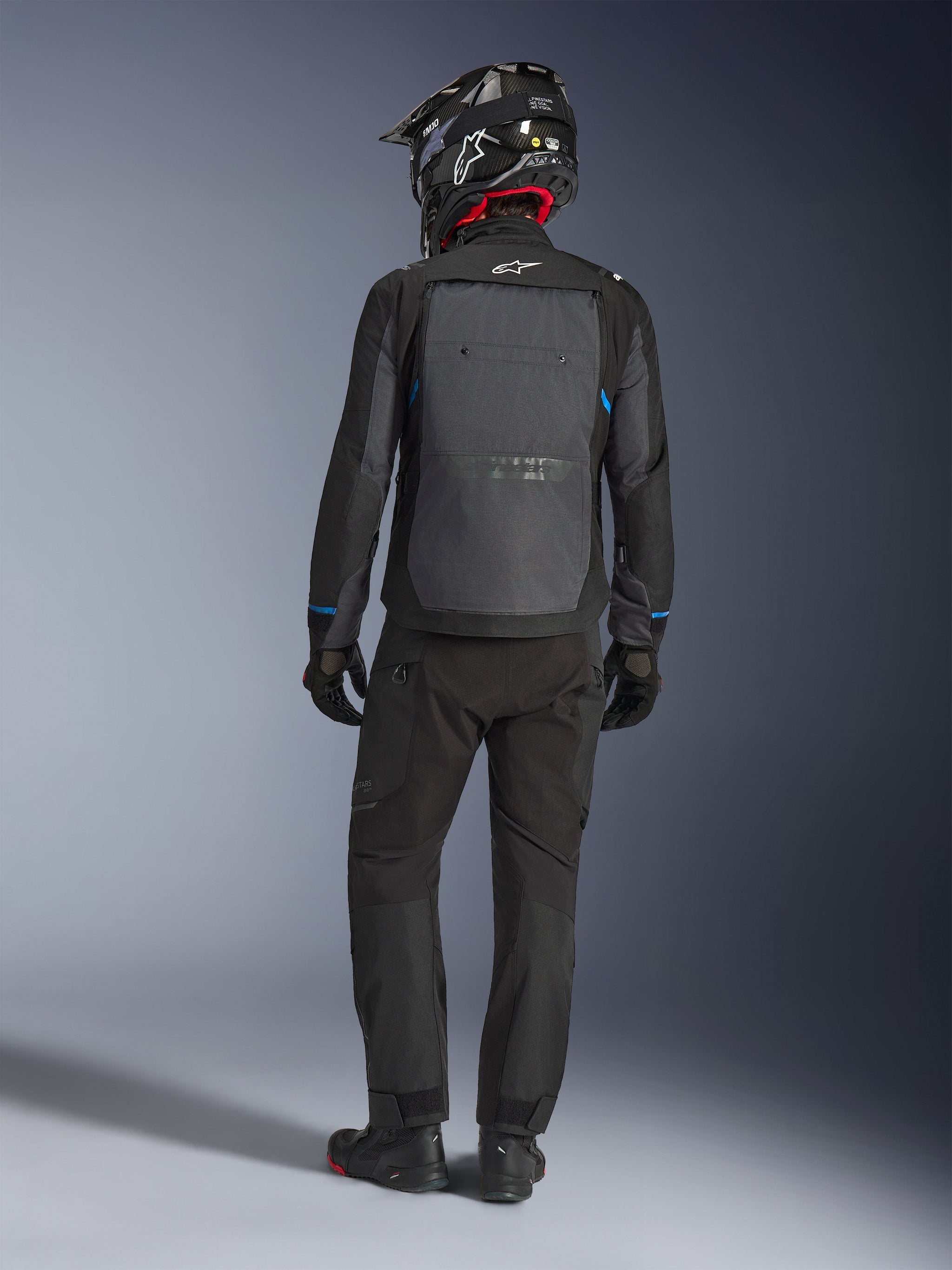 Alpinestars Cusco Drystar® jacka, svart och djupgrå, ryggvy av förare som bär komplett adventure touring-utrustning, stor grå ryggpanel med Alpinestars-logotyp, blå detaljer på ärmarna, bärs med matchande svarta byxor och hjälm.