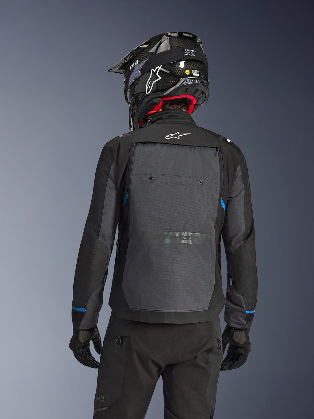 Motorcyklist sedd bakifrån bärande Alpinestars Cusco Drystar®-jacka i Black Deep Gray, med en stor grå ventilationspanel på ryggen och vit logotyp, kombinerat med en svart off-road-hjälm och svart körutrustning för äventyrstouring.