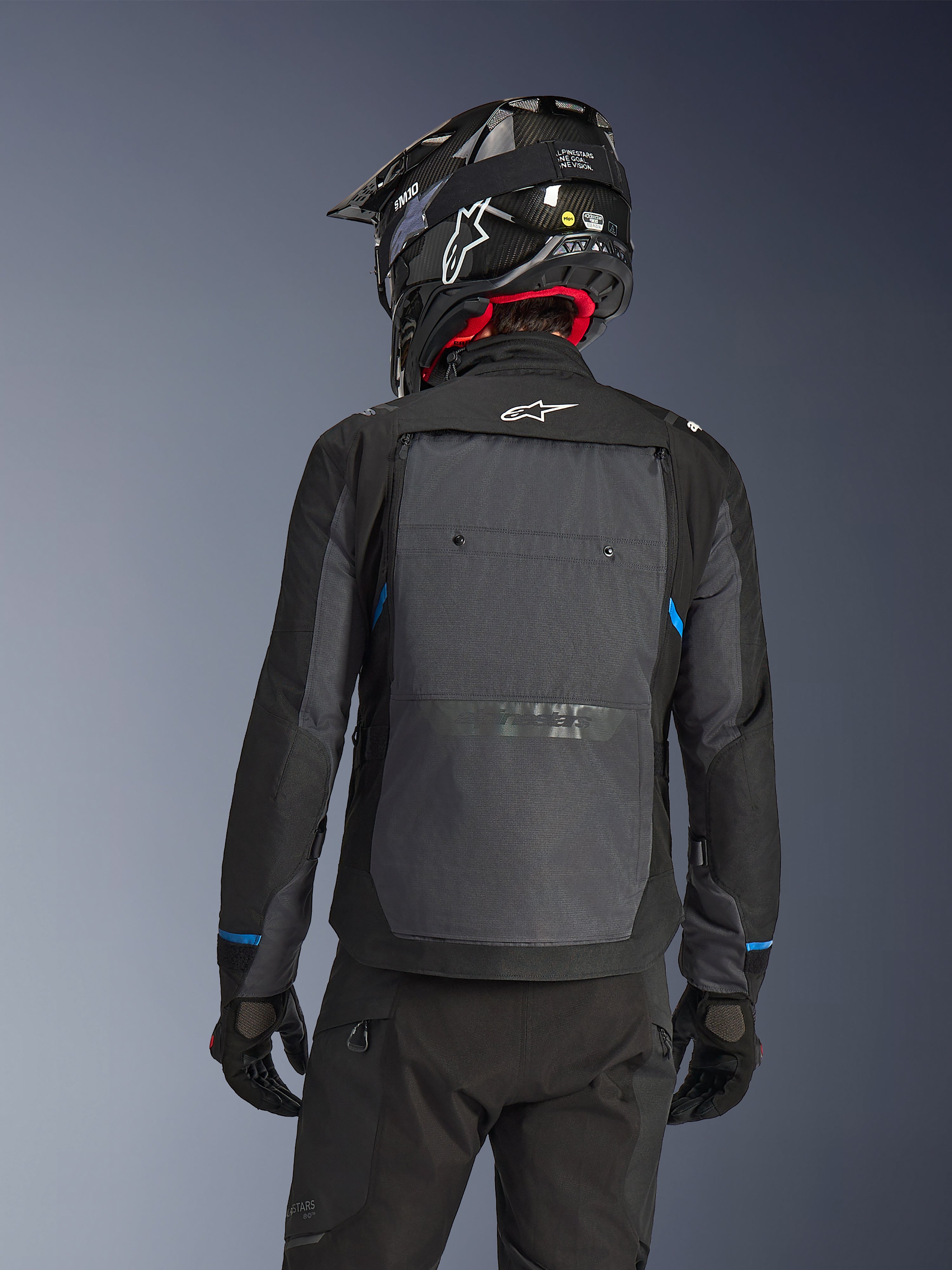 Motorcyklist sedd bakifrån bärande Alpinestars Cusco Drystar®-jacka i Black Deep Gray, med en stor grå ventilationspanel på ryggen och vit logotyp, kombinerat med en svart off-road-hjälm och svart körutrustning för äventyrstouring.