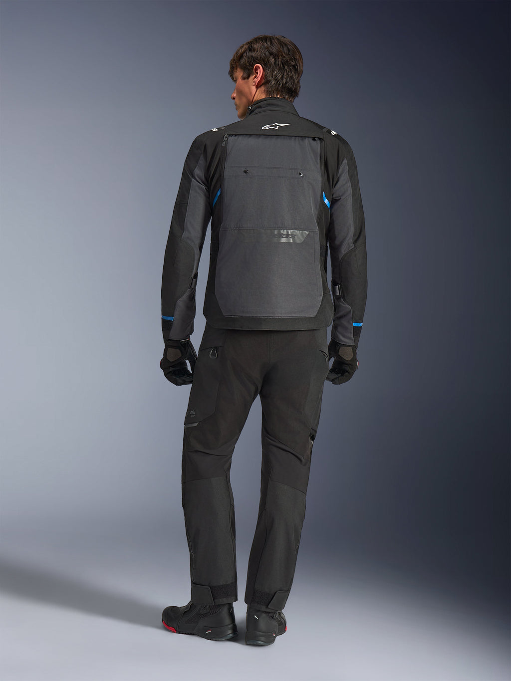 Alpinestars Cusco Drystar®-jacka, svart och mörkgrå med blå detaljer, sedd bakifrån, bärs med matchande svarta äventyrsbyxor och handskar, med en stor grå ryggpanel och vit logotyp på kragen, designad för väg- och äventyrstouring