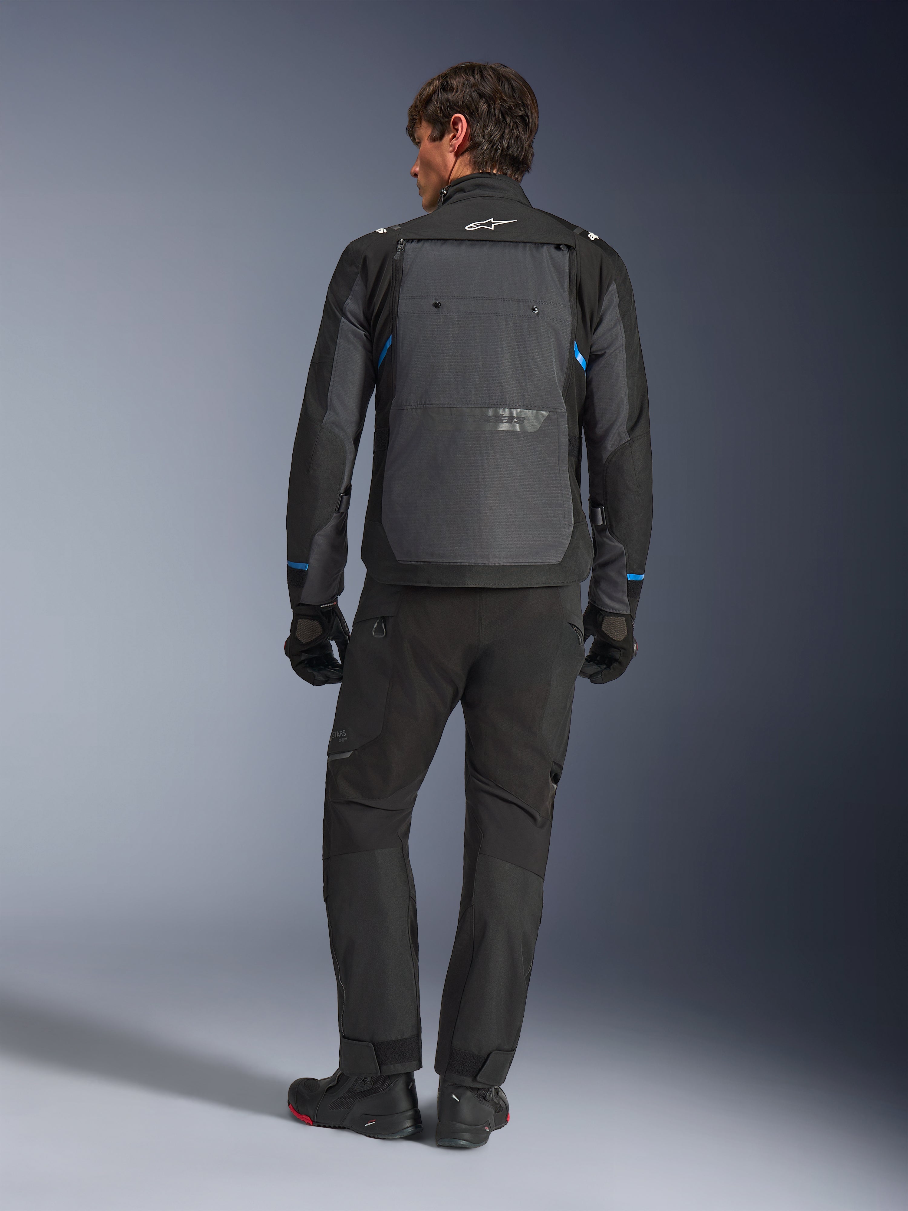Alpinestars Cusco Drystar®-jacka, svart och mörkgrå med blå detaljer, sedd bakifrån, bärs med matchande svarta äventyrsbyxor och handskar, med en stor grå ryggpanel och vit logotyp på kragen, designad för väg- och äventyrstouring
