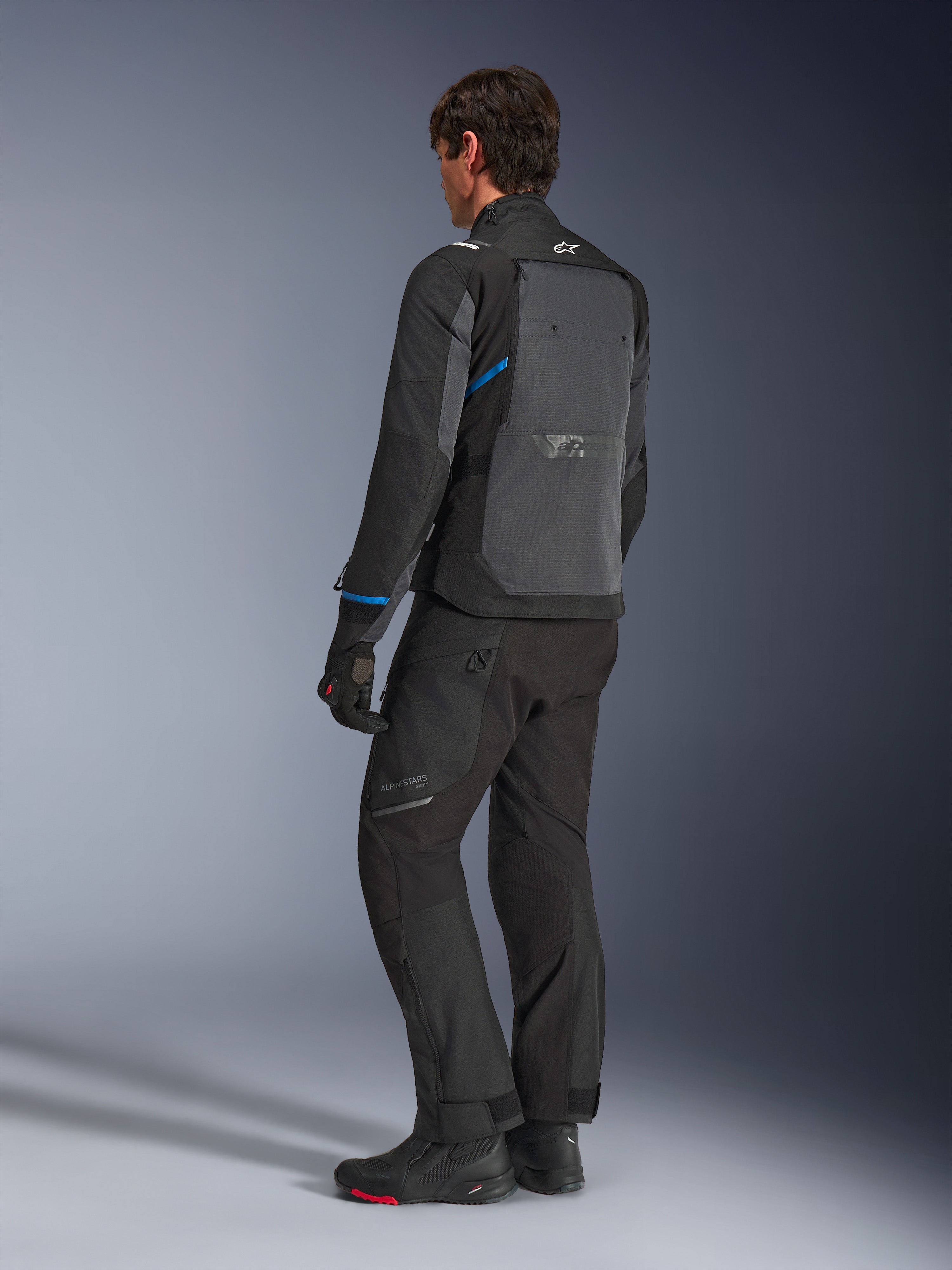 Alpinestars Cusco Drystar® jacka, svart och mörkgrå, trekvartsvy bakifrån som visar en stor ventilationspanel på ryggen och blå detaljer på ärmarna, buren med svarta motorcykelbyxor och stövlar för en komplett touring-outfit.