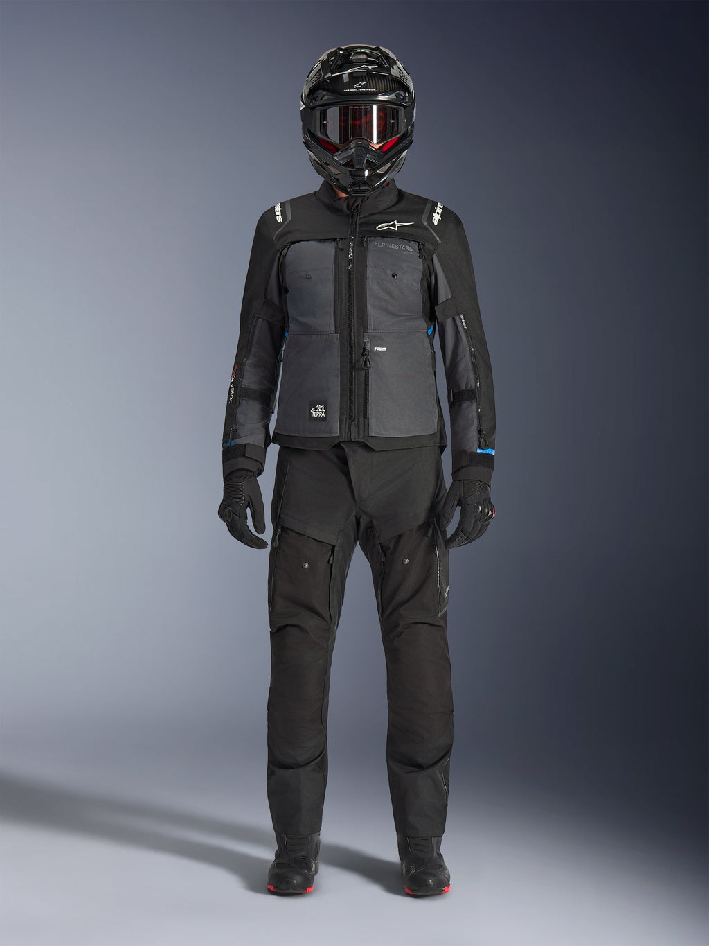 Alpinestars Cusco Drystar®-jacka, svart och mörkgrå adventure touring-jacka, bärs med matchande svarta byxor, handskar och en helhjälm, helkroppsbild framifrån, skyddande motorcykelutrustning