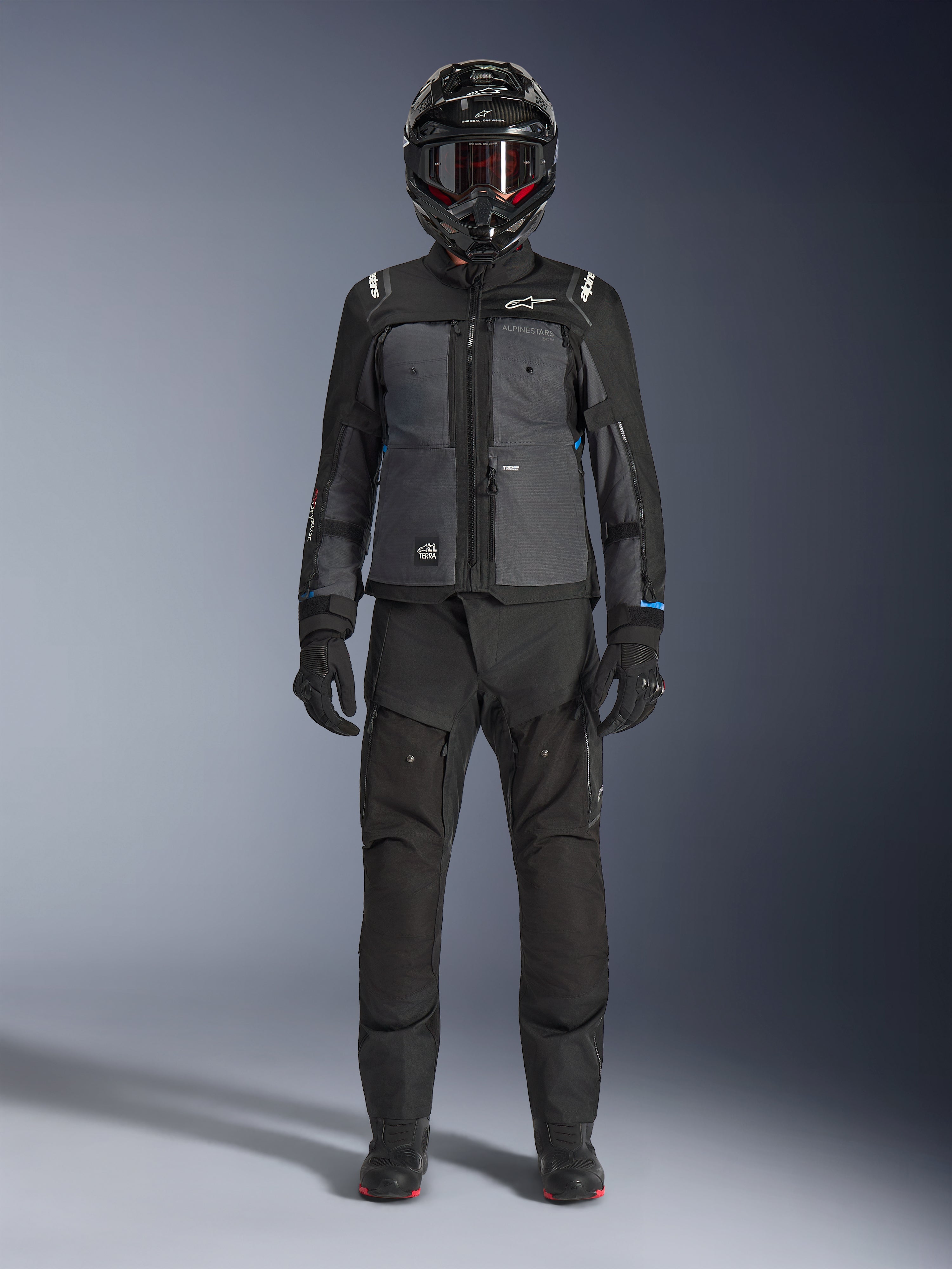 Alpinestars Cusco Drystar®-jacka, svart och mörkgrå adventure touring-jacka, bärs med matchande svarta byxor, handskar och en helhjälm, helkroppsbild framifrån, skyddande motorcykelutrustning
