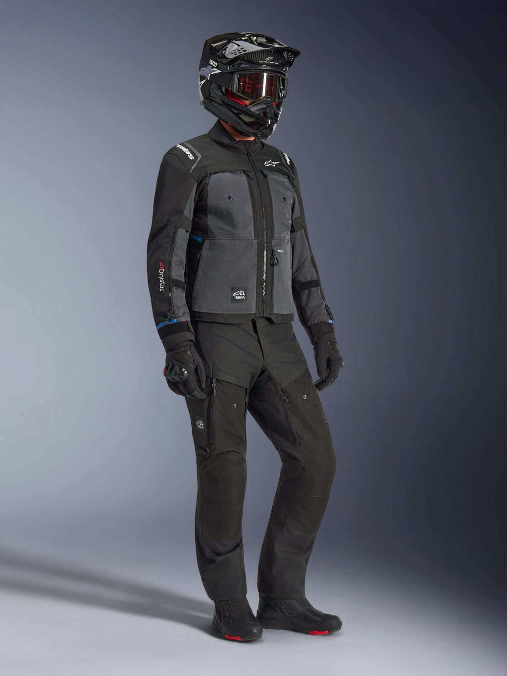 Modell som bär Alpinestars Cusco Drystar®-jacka i Black och Deep Gray, kombinerat med komplett motorcykelutrustning inklusive en svart hjälm, matchande tekniska byxor, handskar och stövlar, med ripstop- och softshell-konstruktion för väg- och äventyrstouring