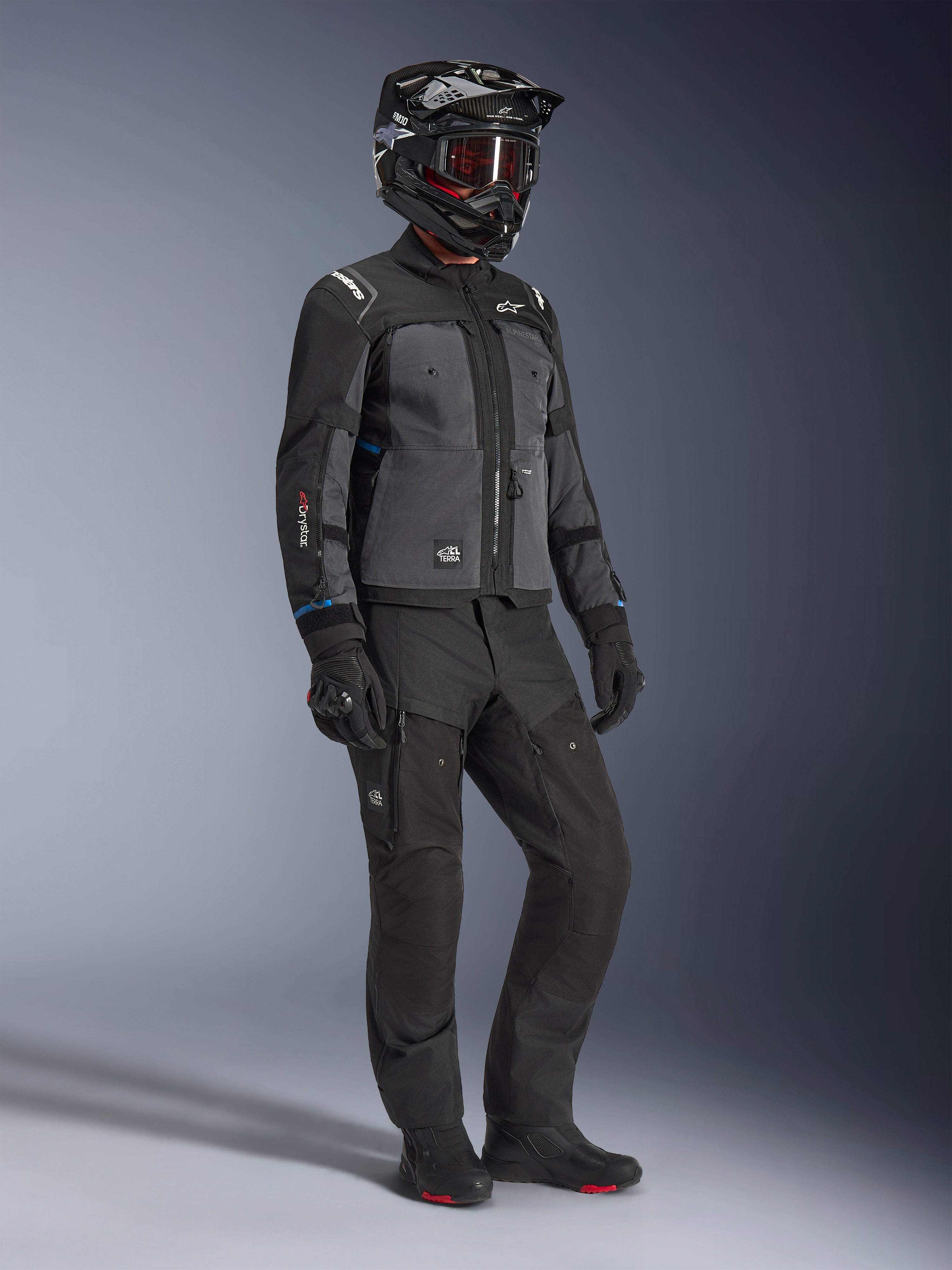 Modell som bär Alpinestars Cusco Drystar®-jacka i Black och Deep Gray, kombinerat med komplett motorcykelutrustning inklusive en svart hjälm, matchande tekniska byxor, handskar och stövlar, med ripstop- och softshell-konstruktion för väg- och äventyrstouring
