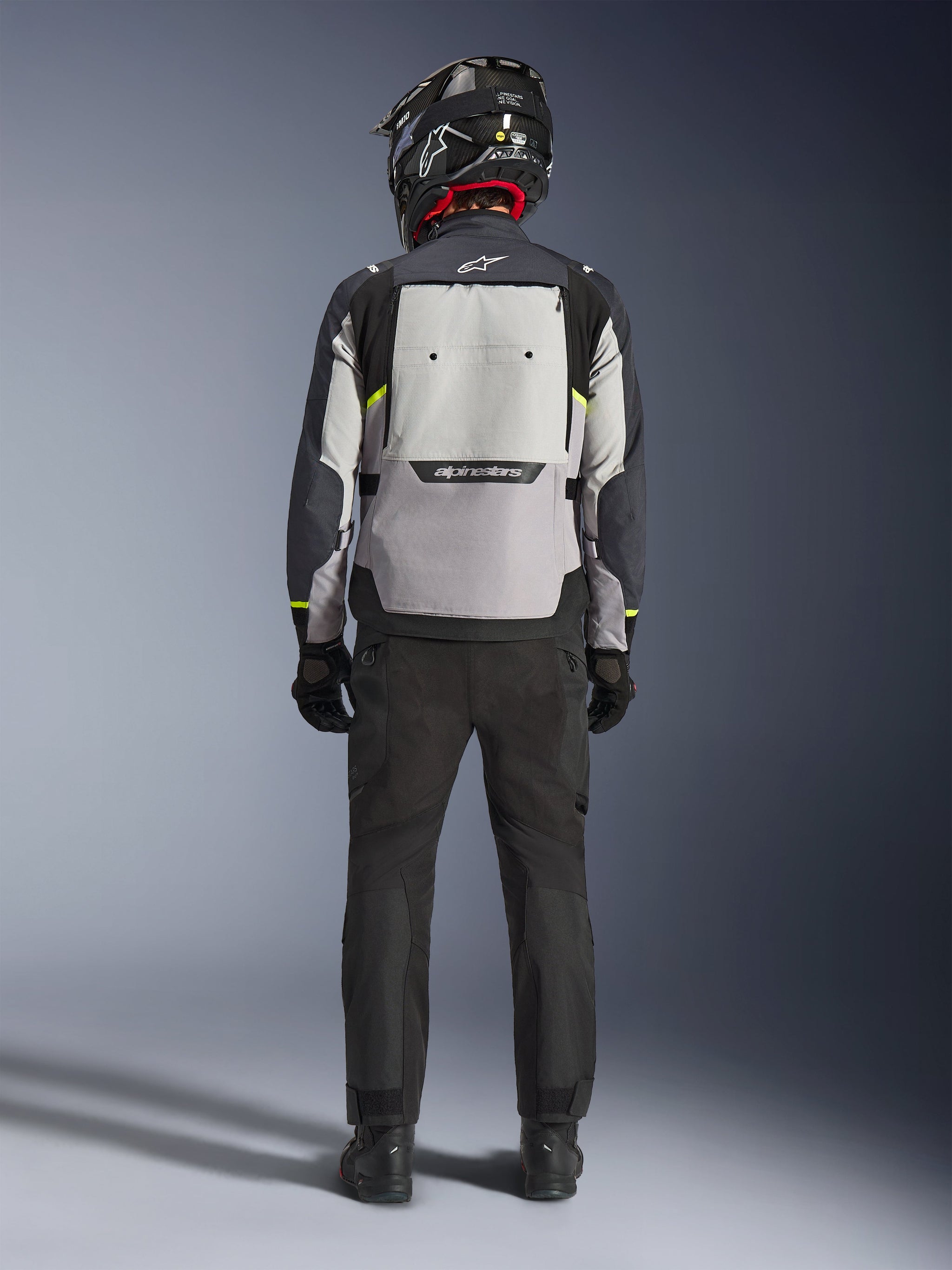 Motorcykelförare sedd bakifrån iklädd Alpinestars Cusco Drystar®-jacka i Ice Gray, Mid Gray och Deep Gray, med en stor ventilationspanel på ryggen och Alpinestars-logotyp, kombinerad med svarta körbyxor och en svart hjälm, adventure touring-motorcykelkläder.