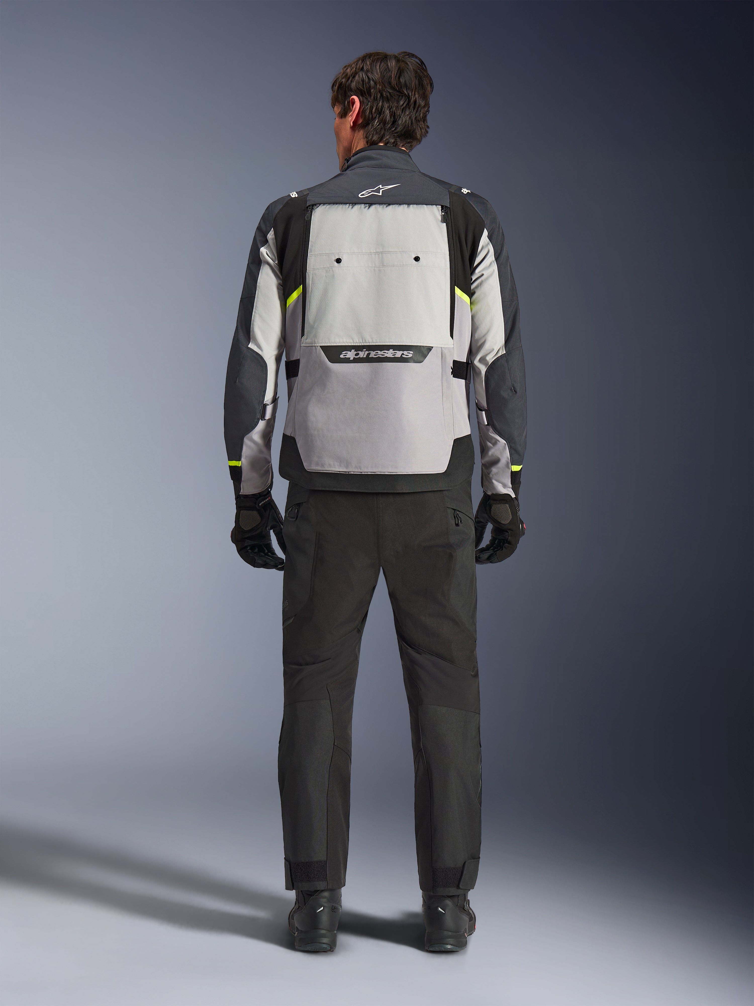 Vy bakifrån av en förare som bär Alpinestars Cusco Drystar®-jacka i Ice Gray, Mid Gray och Deep Gray, matchad med svarta motorcykelbyxor och handskar, vilket framhäver den stora ventilationspanelen på ryggen och Alpinestars logotyp.