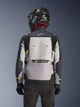 Alpinestars Cusco Drystar®-jacka, baksida, Ice Gray, Mid Gray och Deep Gray, med stor bakre ventilationspanel och högvisibla gula detaljer, bärs med en svart äventyrshjälm och svarta byxor.