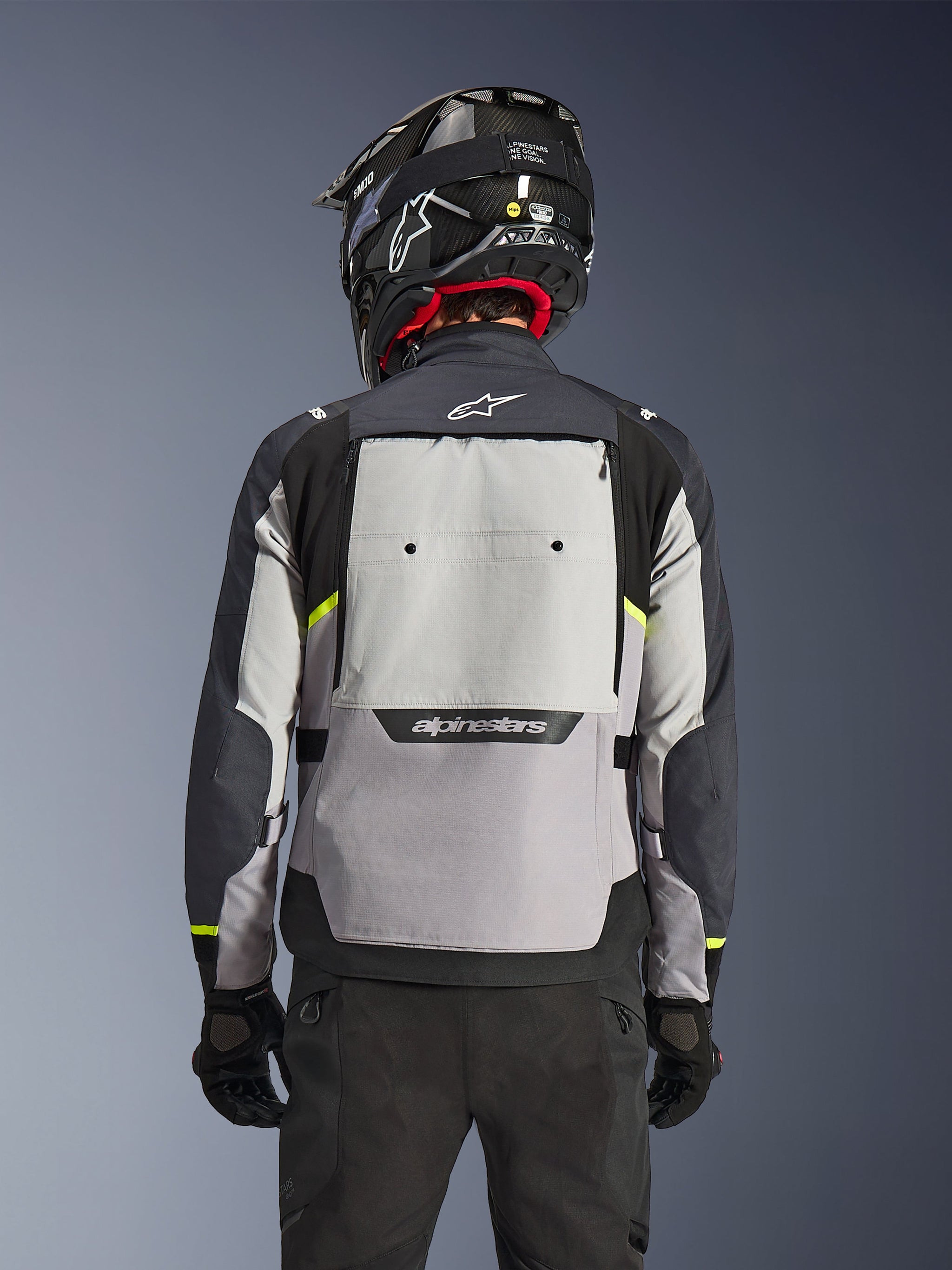 Alpinestars Cusco Drystar®-jacka, baksida, Ice Gray, Mid Gray och Deep Gray, med stor bakre ventilationspanel och högvisibla gula detaljer, bärs med en svart äventyrshjälm och svarta byxor.