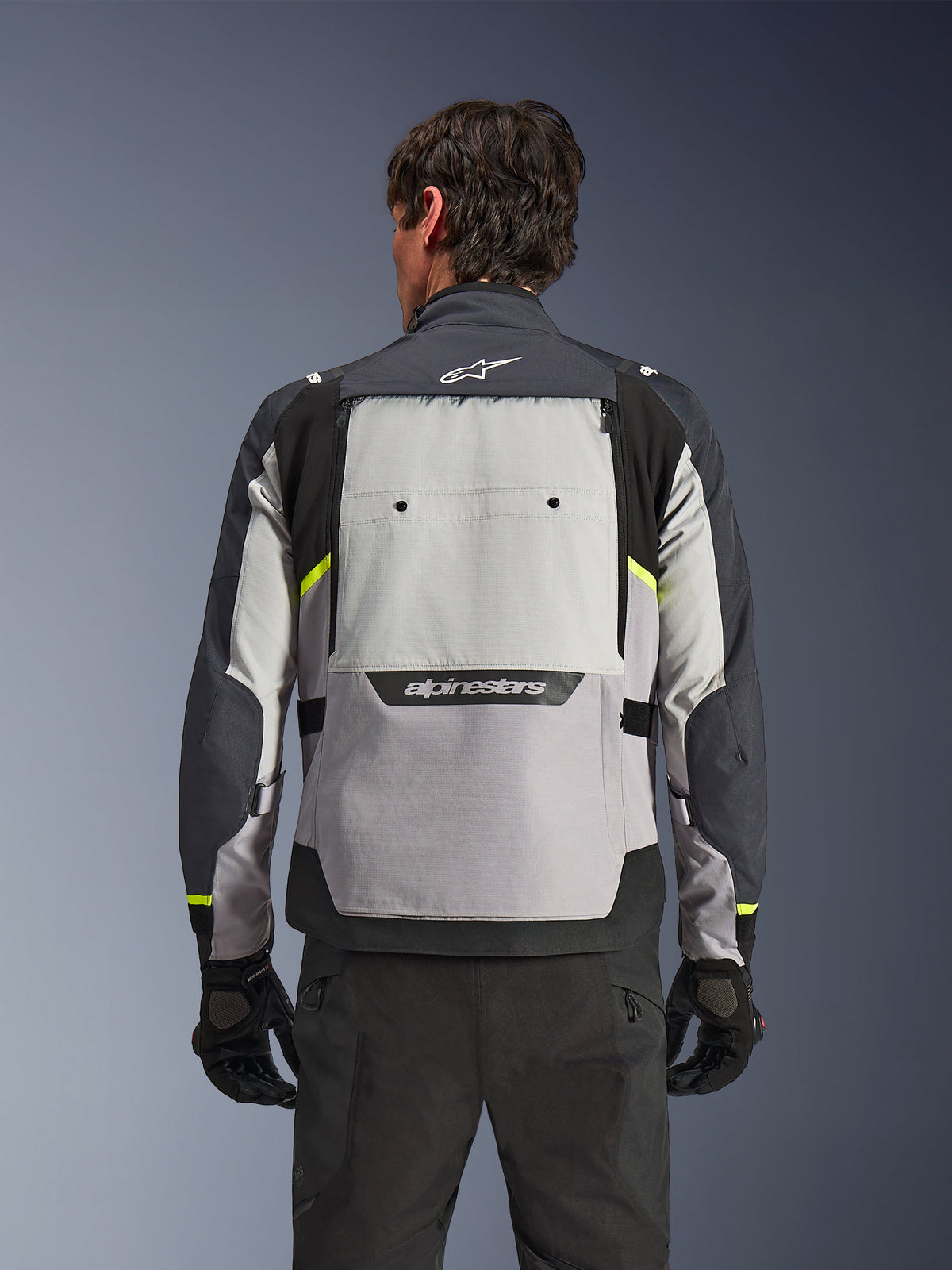 Bakifrån sedd förare som bär Alpinestars Cusco Drystar®-jacka i färgerna Ice Gray, Mid Gray och Deep Gray, med neongula accenter, en stor ventilationspanel på ryggen och tydlig Alpinestars-logotyp, matchad med svarta körbyxor och handskar.