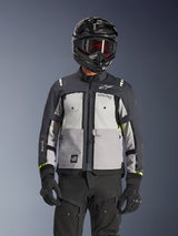 Alpinestars Cusco Drystar® jacka, äventyrsjacka, isgrå, mellangrå och djupgrå, med paneler i flera grå nyanser, ventilationsöppningar med dragkedja på bröstet och Alpinestars-logotyper, bärs av en förare med svart hjälm och goggles.