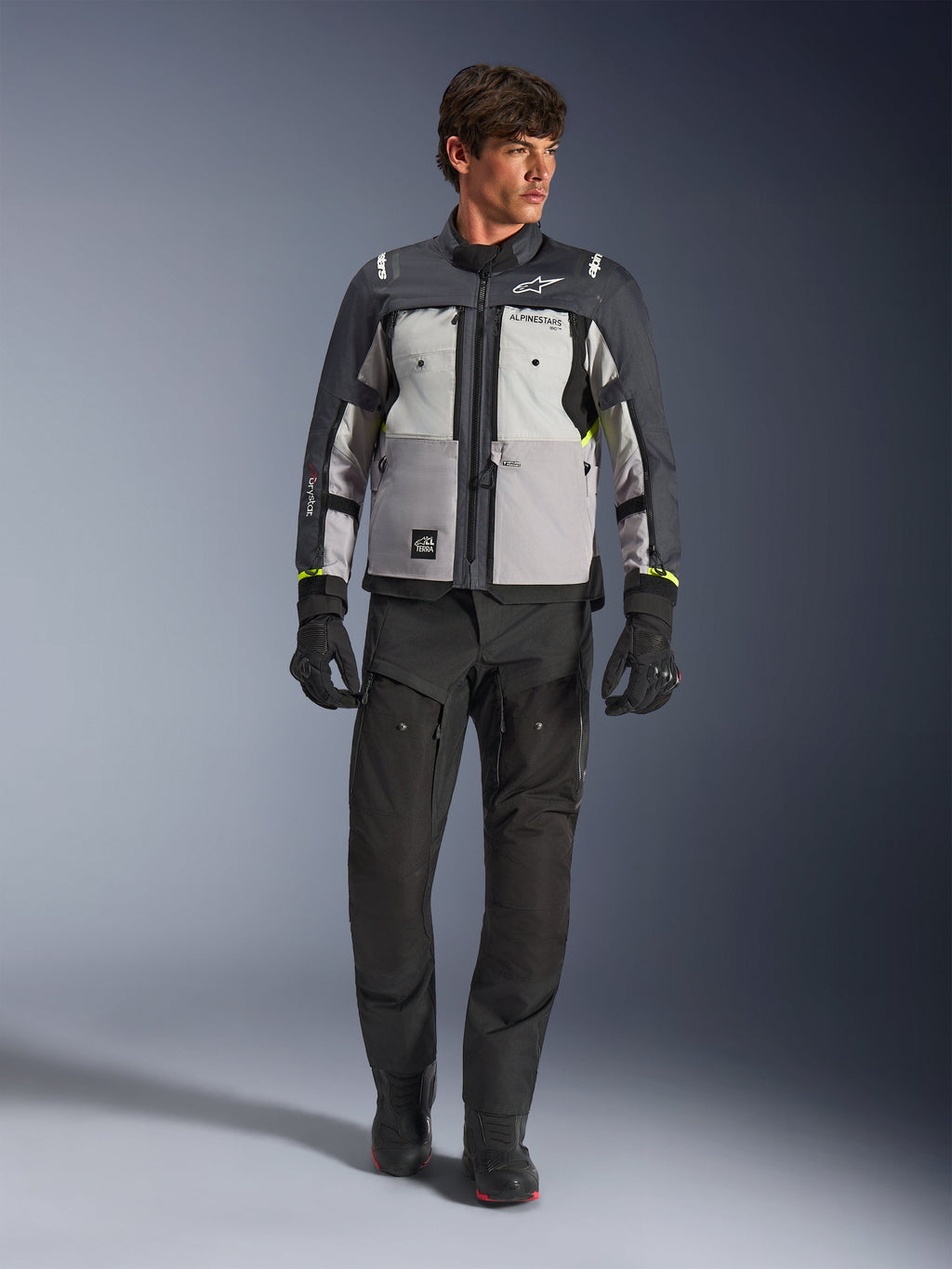 Alpinestars Cusco Drystar® jacka i Ice Gray, Mid Gray och Deep Gray, bärs med svarta adventure-motorcykelbyxor och handskar, textil adventure touring-jacka med grå paneler i flera toner och neongula detaljer, studiobild på manlig modell.