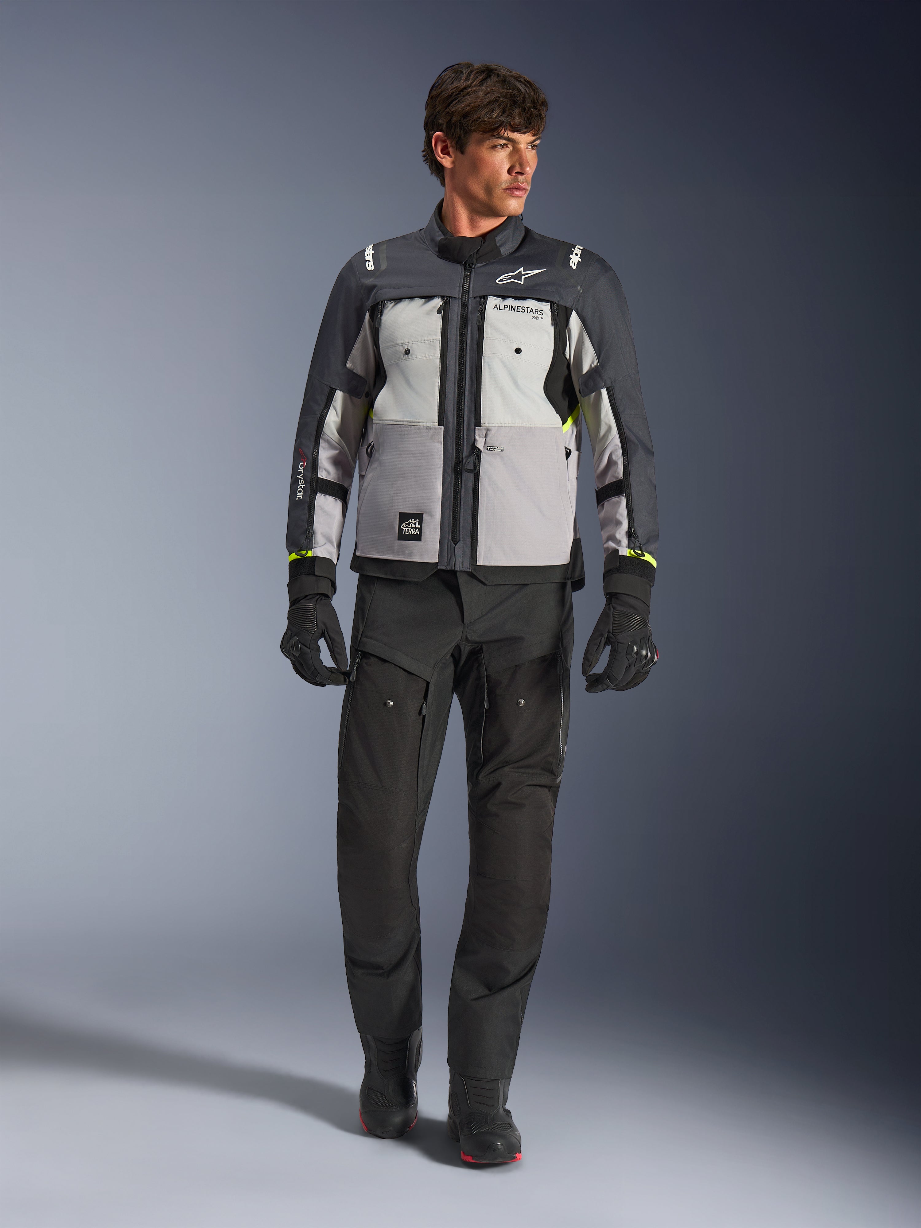 Alpinestars Cusco Drystar® jacka i Ice Gray, Mid Gray och Deep Gray, bärs med svarta adventure-motorcykelbyxor och handskar, textil adventure touring-jacka med grå paneler i flera toner och neongula detaljer, studiobild på manlig modell.