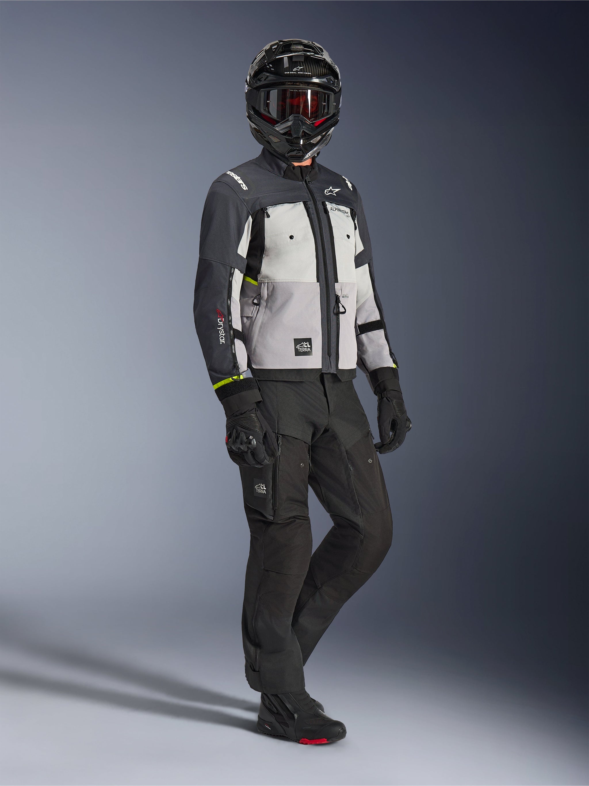 Alpinestars Cusco Drystar® jacka i Ice Gray Mid Gray Deep Gray, helkroppsbild av en förare iklädd komplett äventyrsutrustning inklusive en svart hjälm med röda goggles, matchande svarta byxor och handskar, textilmotorcykeljacka med direktventilationspaneler och vattentätt membran