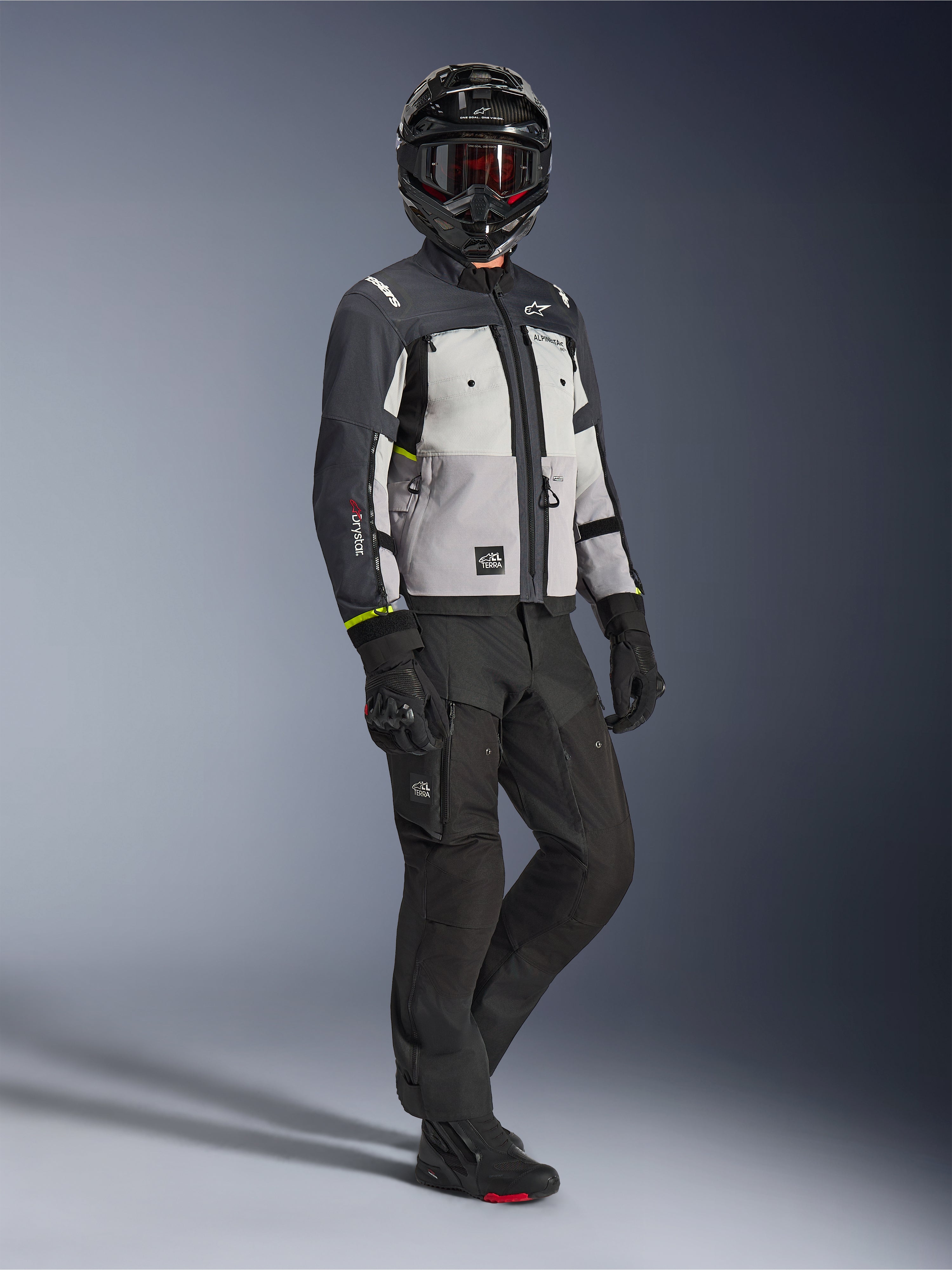 Alpinestars Cusco Drystar® jacka i Ice Gray Mid Gray Deep Gray, helkroppsbild av en förare iklädd komplett äventyrsutrustning inklusive en svart hjälm med röda goggles, matchande svarta byxor och handskar, textilmotorcykeljacka med direktventilationspaneler och vattentätt membran