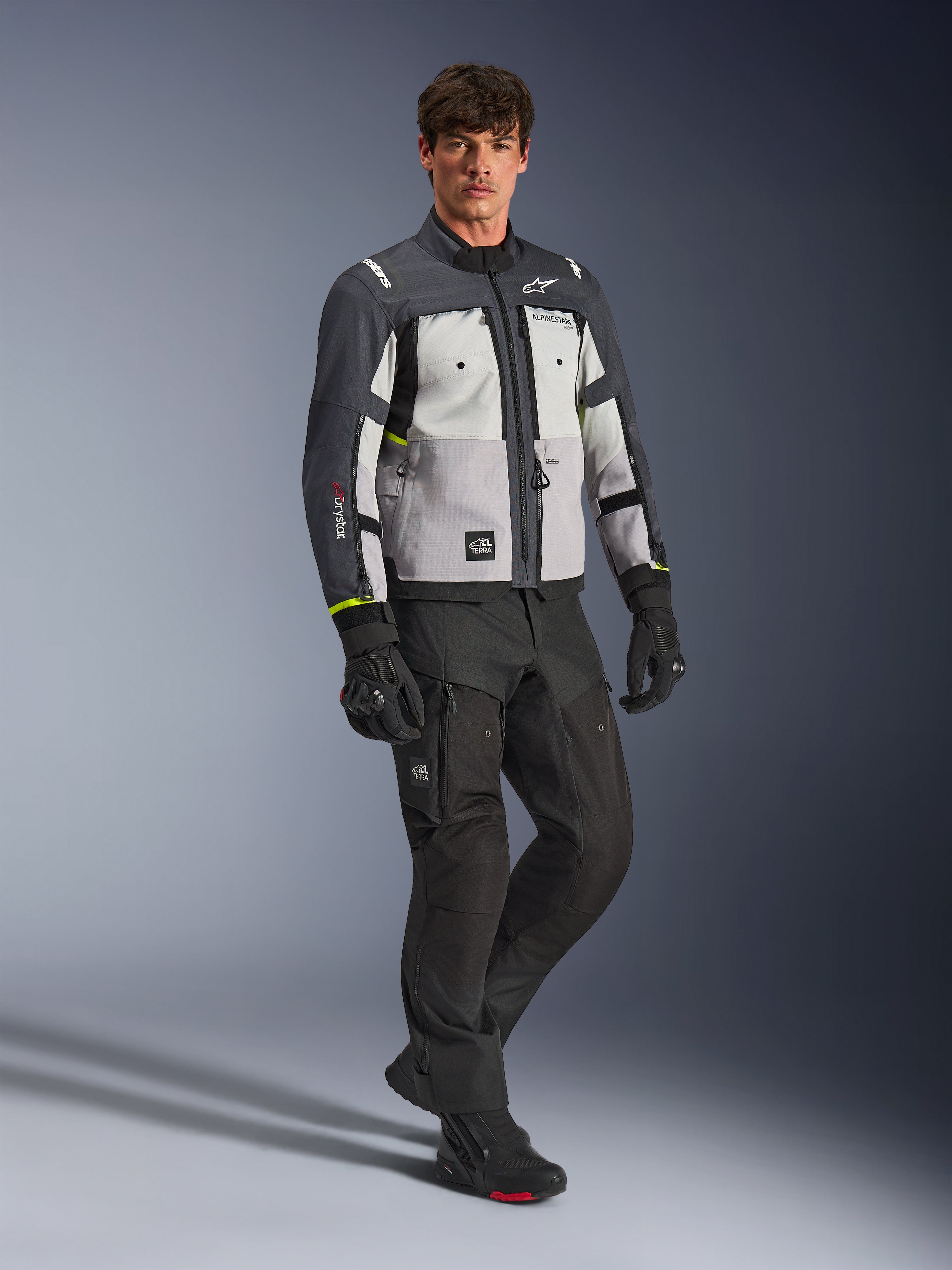 Alpinestars Cusco Drystar®-jacka, äventyrsjacka för motorcykel, Ice Gray, Mid Gray och Deep Gray, bärs av en modell med svarta byxor och handskar, grå paneler i flera nyanser med hållbar textilkonstruktion, med ventilationsöppningar med dragkedja och Alpinestars-logotyp, professionell studiofotografering för väg- och äventyrsutrustning.
