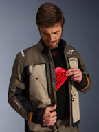 Alpinestars Cusco Drystar®-jacka, Walnut Aluminum Black, bärs av en man som demonstrerar bröstskyddsfickan med ett rött skyddsinlägg, textilmotorcykeljacka med ventilationsöppningar med dragkedja och märkesdetaljer för adventure touring.