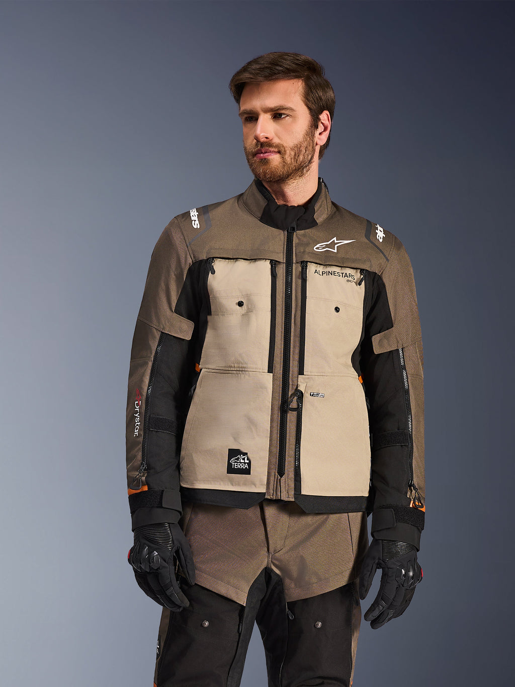 Alpinestars Cusco Drystar®-jacka, äventyrsmotorcykeljacka, Walnut Aluminum Black, bärs av en modell med svarta handskar, med beige bröstpaneler och svarta ärmar med Alpinestars-logotyp och All Terra-märke, skyddande touringutrustning för alla väder