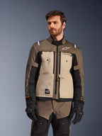 Alpinestars Cusco Drystar®-jacka, äventyrsmotorcykeljacka, Walnut Aluminum Black, bärs av en modell med svarta handskar, med beige bröstpaneler och svarta ärmar med Alpinestars-logotyp och All Terra-märke, skyddande touringutrustning för alla väder
