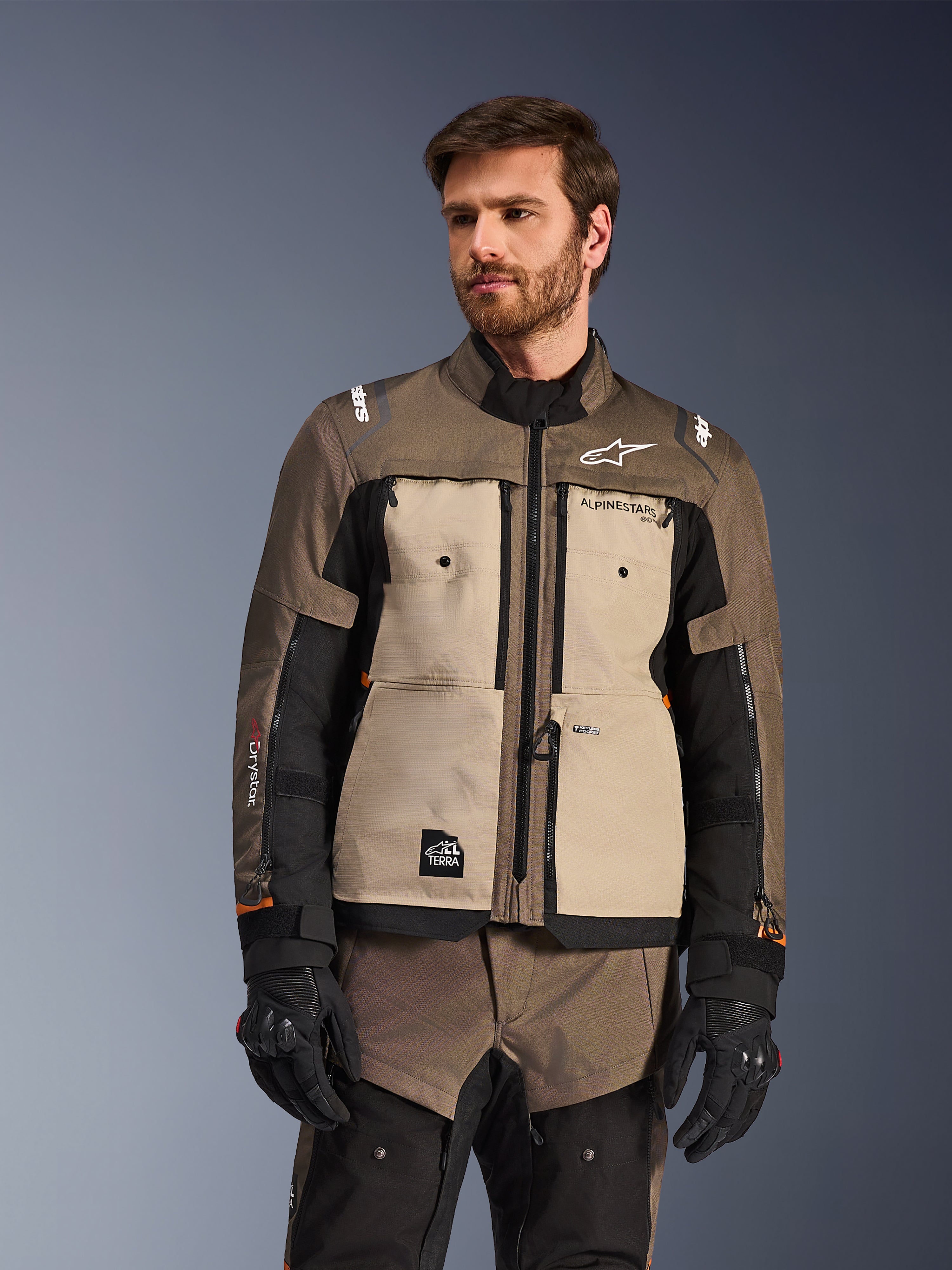 Alpinestars Cusco Drystar®-jacka, äventyrsmotorcykeljacka, Walnut Aluminum Black, bärs av en modell med svarta handskar, med beige bröstpaneler och svarta ärmar med Alpinestars-logotyp och All Terra-märke, skyddande touringutrustning för alla väder