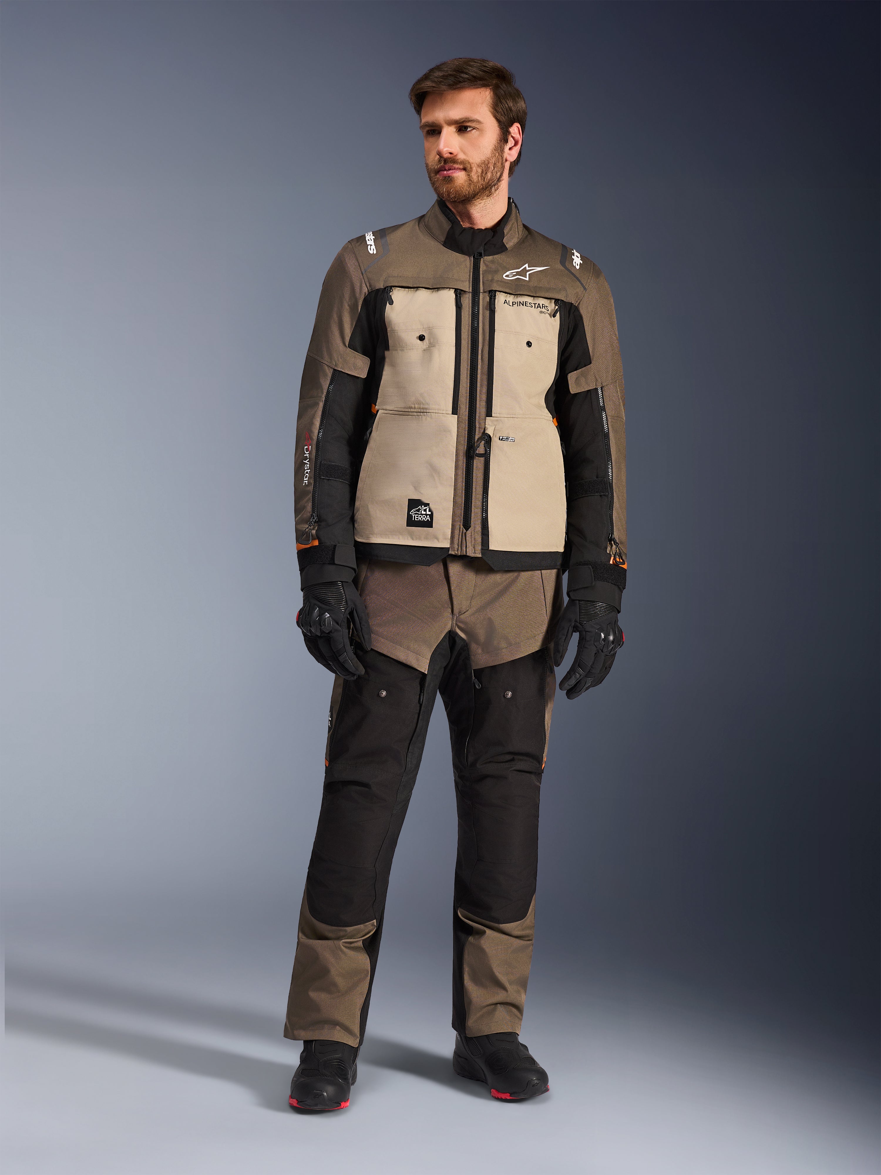 Alpinestars Cusco Drystar®-jacka i Walnut Aluminum Black, bärs av en manlig förare med matchande adventure touring-byxor, svarta handskar och stövlar, med beigea bröstpaneler, svarta ärmar med subtila orangea detaljer och funktionella ventilationsdragkedjor för landsvägskörning.