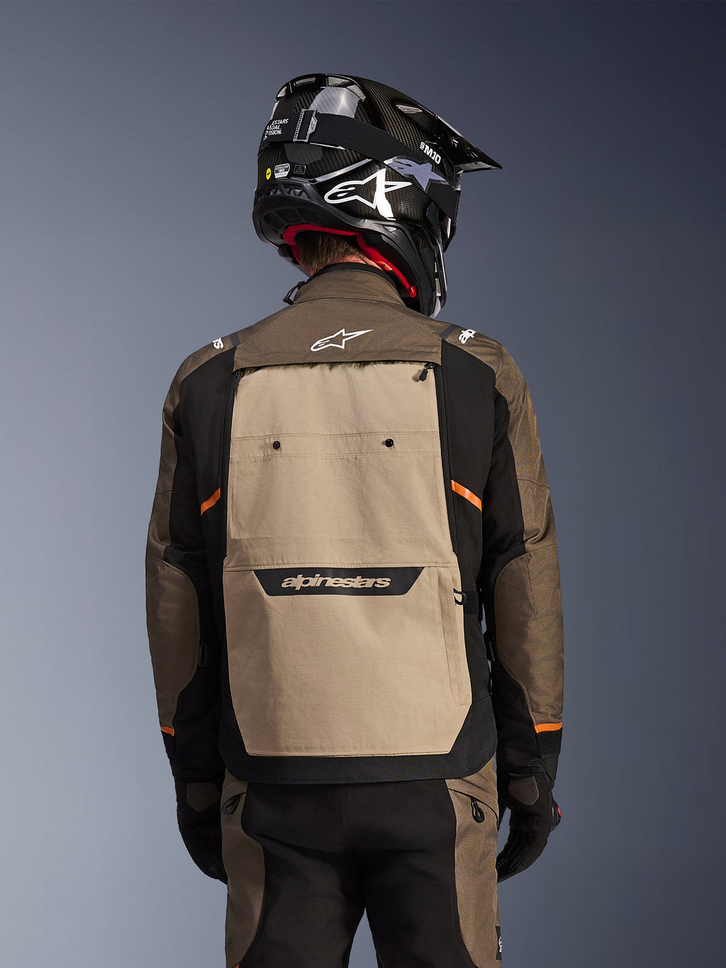 Baksida av Alpinestars Cusco Drystar®-jacka i Walnut Aluminum Black, adventure touring-motorcykeljacka med en stor beige ryggpanel med Alpinestars-logotyp, svarta ärmar med orangea detaljer, visas med en svart hjälm i kolfiberstil.