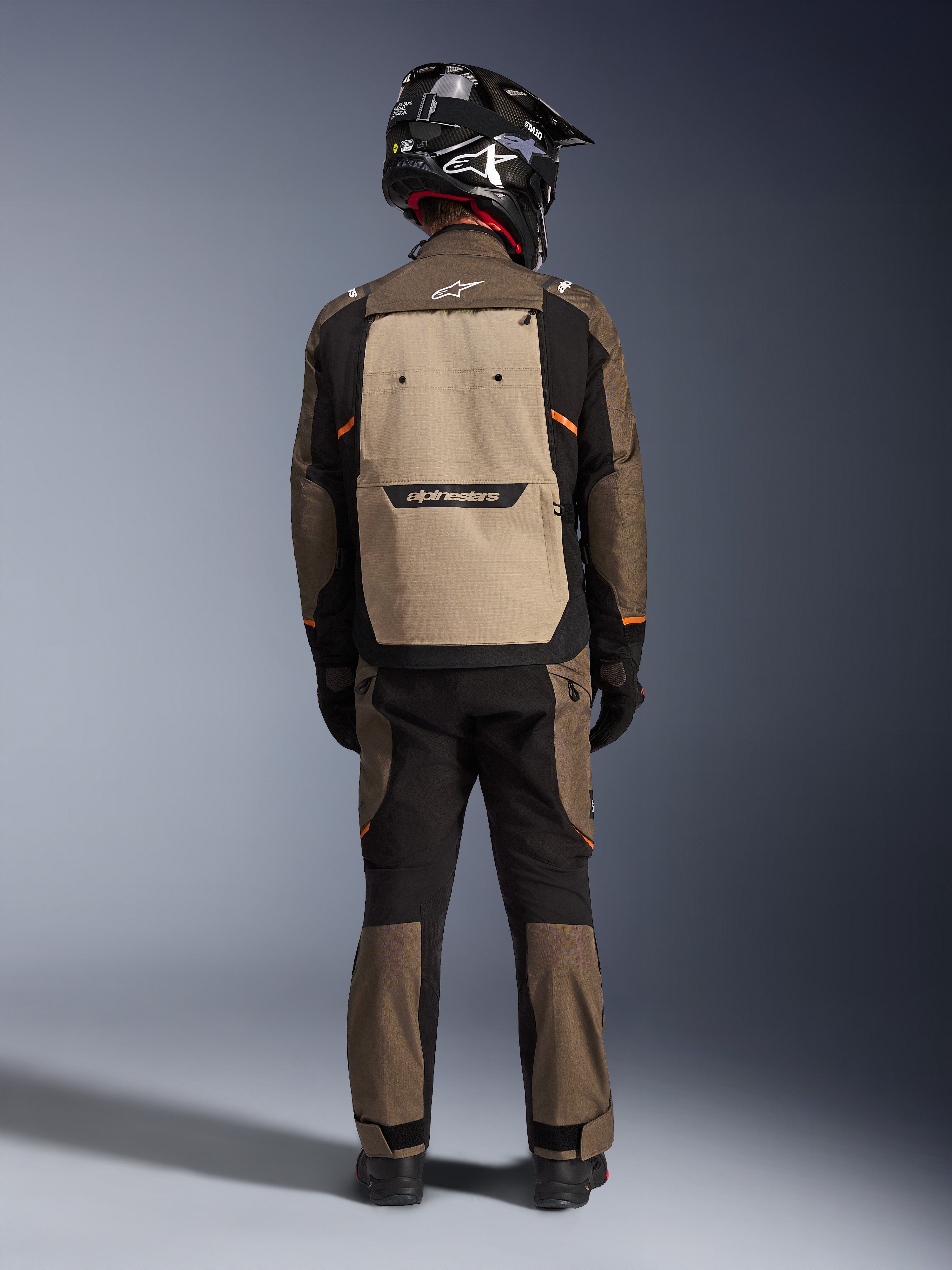 Förare sedd bakifrån i Alpinestars Cusco Drystar®-jacka i Walnut Aluminum Black, med en stor aluminiumfärgad ryggficka och orangea detaljer, matchande adventure-byxor, svart offroad-hjälm och handskar, slitstark MC-utrustning i textil för touring.