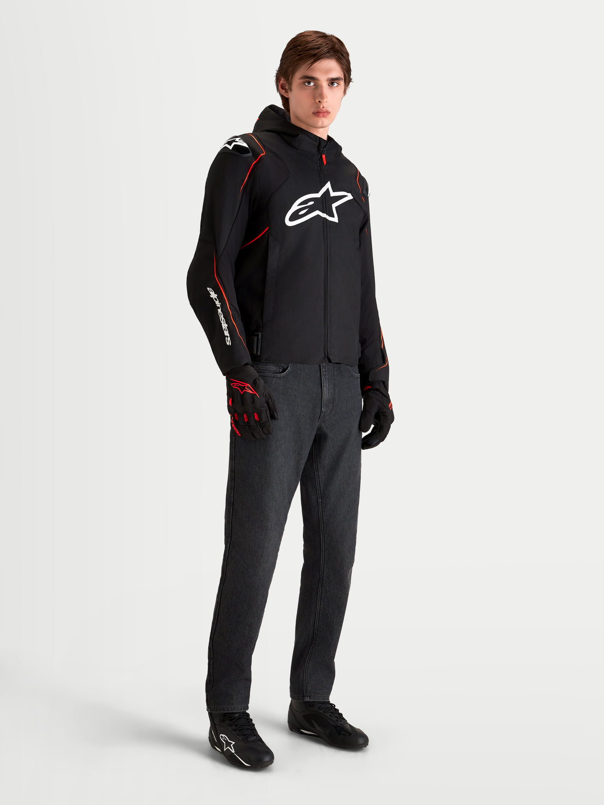 Alpinestars Zaca V 2 Waterproof jacka, svart och röd motorcykelsportjacka för herr med huva och vit bröstlogotyp, bärs av en manlig modell med matchande körhandskar och mörka jeans