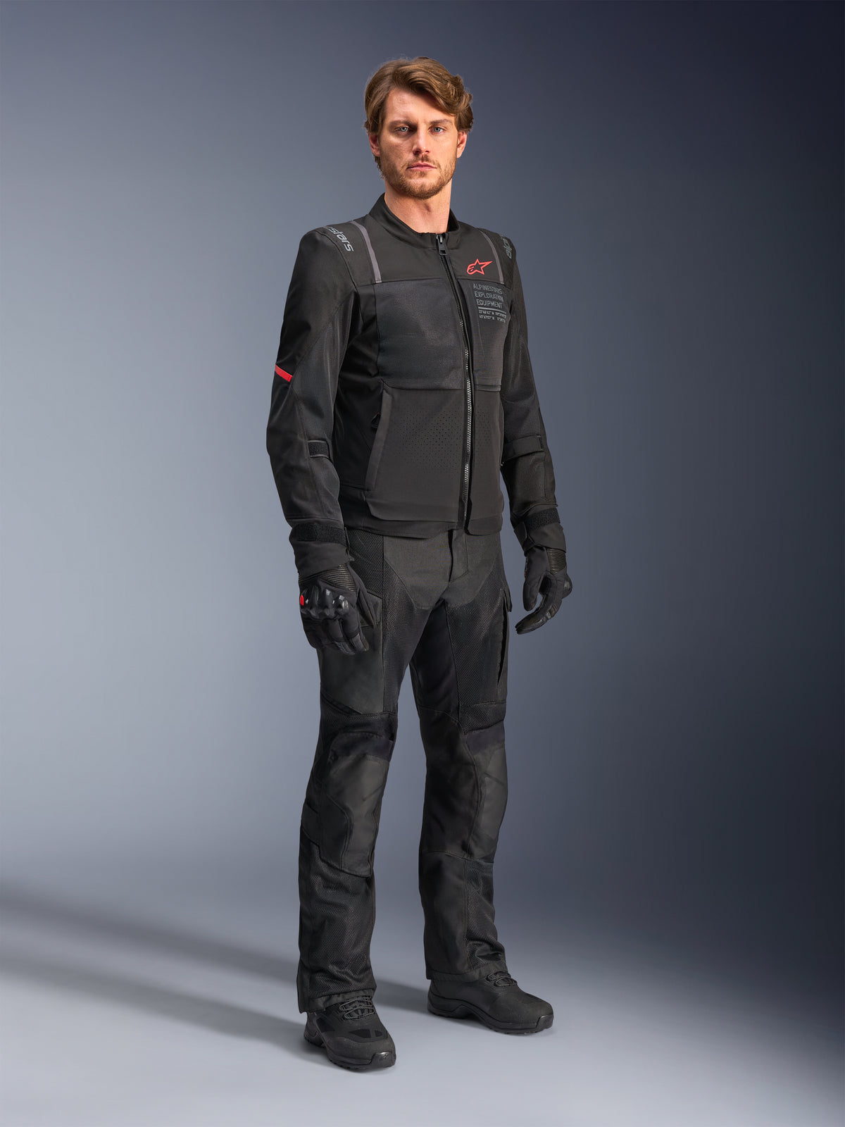 Male model wearing the Alpinestars ST-2 Air Jacket in black, featuring a red star logo and 'Alpinestars Exploration Equipment' branding, paired with matching black riding pants, gloves, and boots for a modern summer touring outfit.