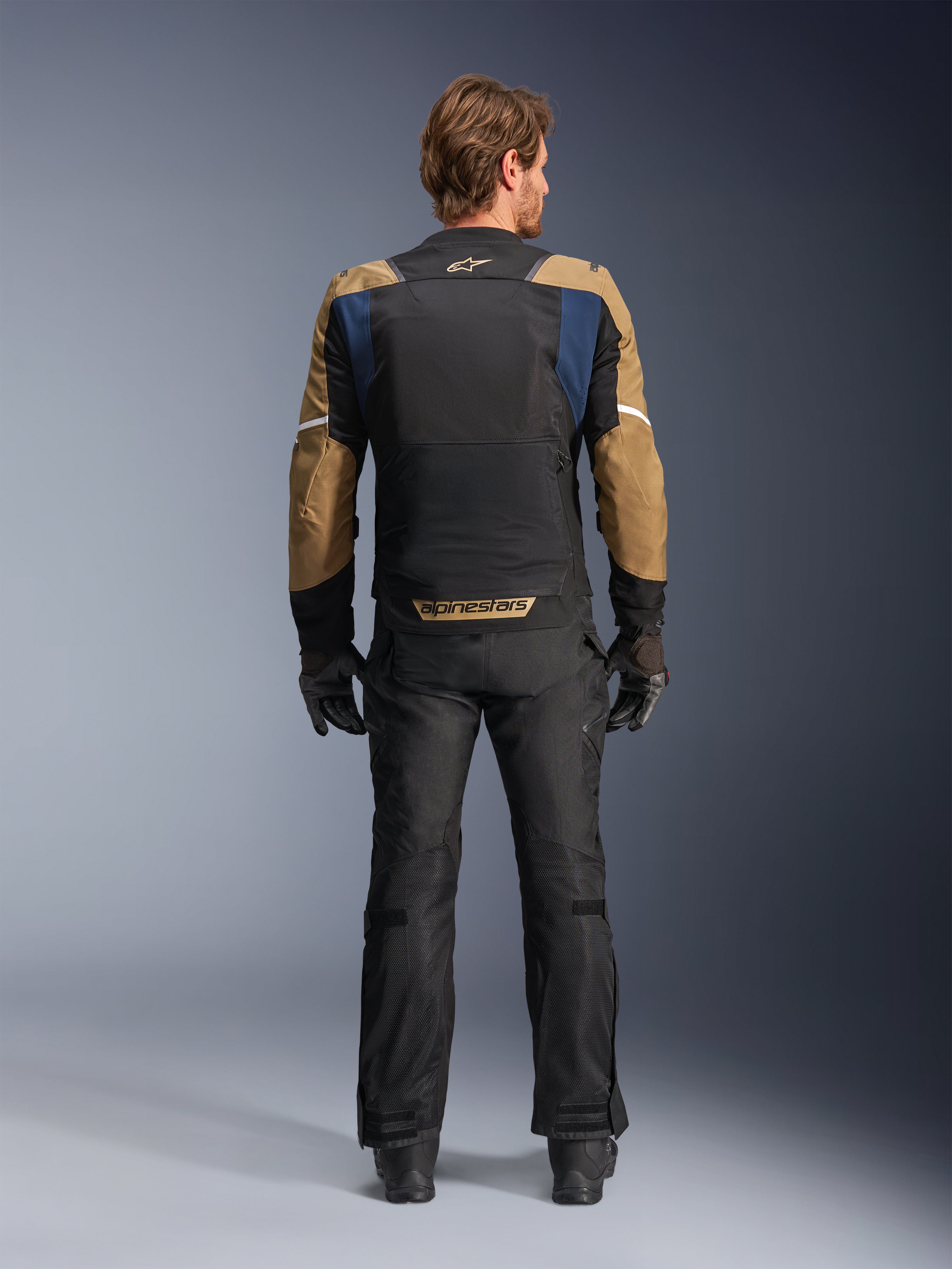 Alpinestars ST-2 Air-jacka i Deep Blue, Dark Khaki och svart, sedd bakifrån på modell som visar stor mesh-ryggpanel och khaki-färgade ärmar med vita ränder, kombinerad med svarta tekniska motorcykelbyxor och handskar för adventure touring.