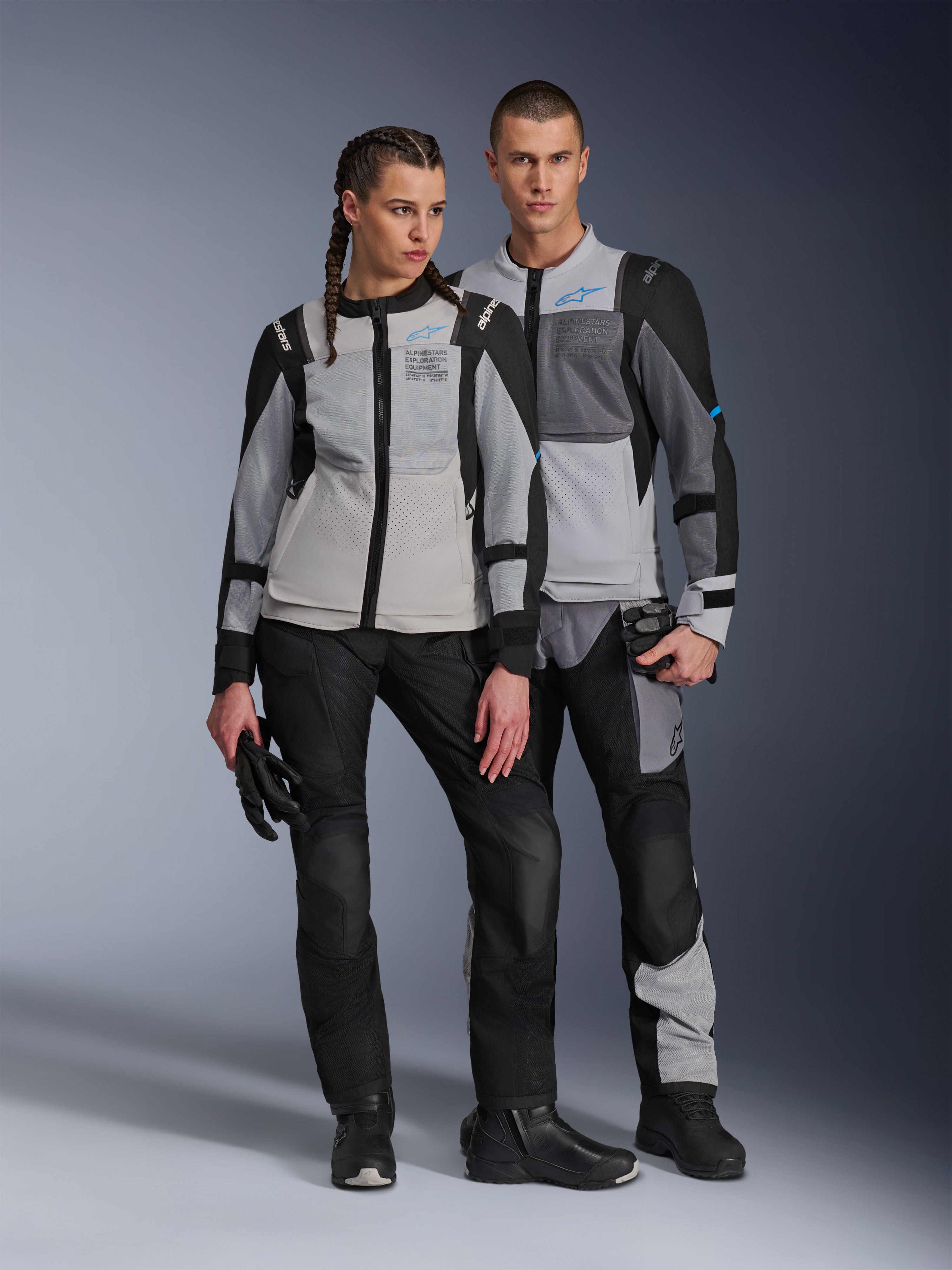 Manlig och kvinnlig modell bär Alpinestars ST-2 Air-jackor i Ice Gray, Dark Gray och Black, teknisk motorcykelutrustning för sommarturer med stora meshpaneler och exploration equipment-märkning, matchat med svarta körbyxor, handskar och stövlar.
