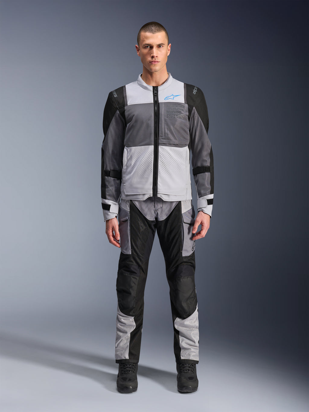 Alpinestars ST-2 Air jacka, touringjacka för motorcykel, Ice Gray, mörkgrå och svart, bärs av en modell med matchande adventure-byxor, stora meshpaneler, blå Alpinestars-logotyp på bröstet och Alpinestars Exploration Equipment-märkning