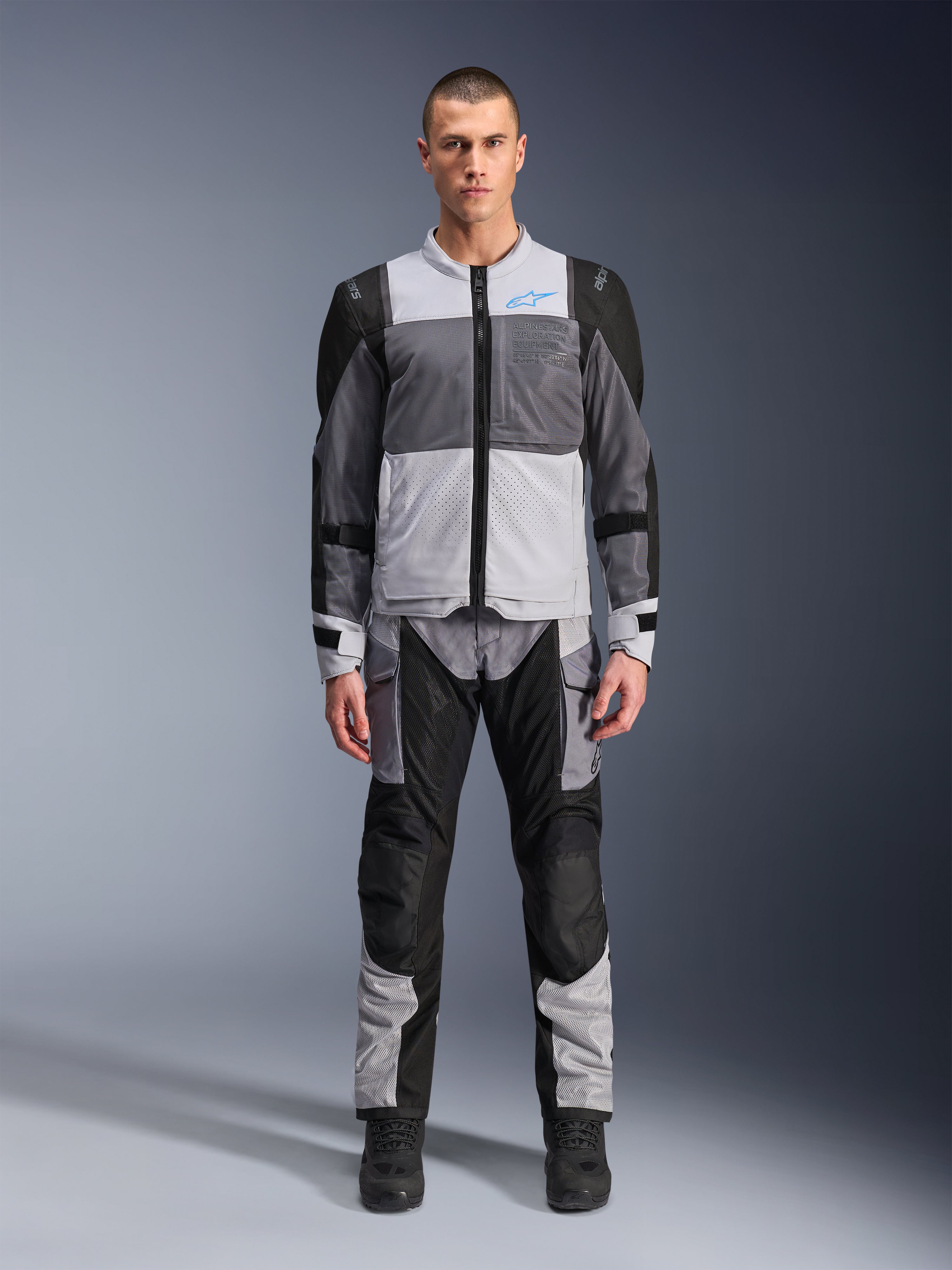 Alpinestars ST-2 Air jacka, touringjacka för motorcykel, Ice Gray, mörkgrå och svart, bärs av en modell med matchande adventure-byxor, stora meshpaneler, blå Alpinestars-logotyp på bröstet och Alpinestars Exploration Equipment-märkning