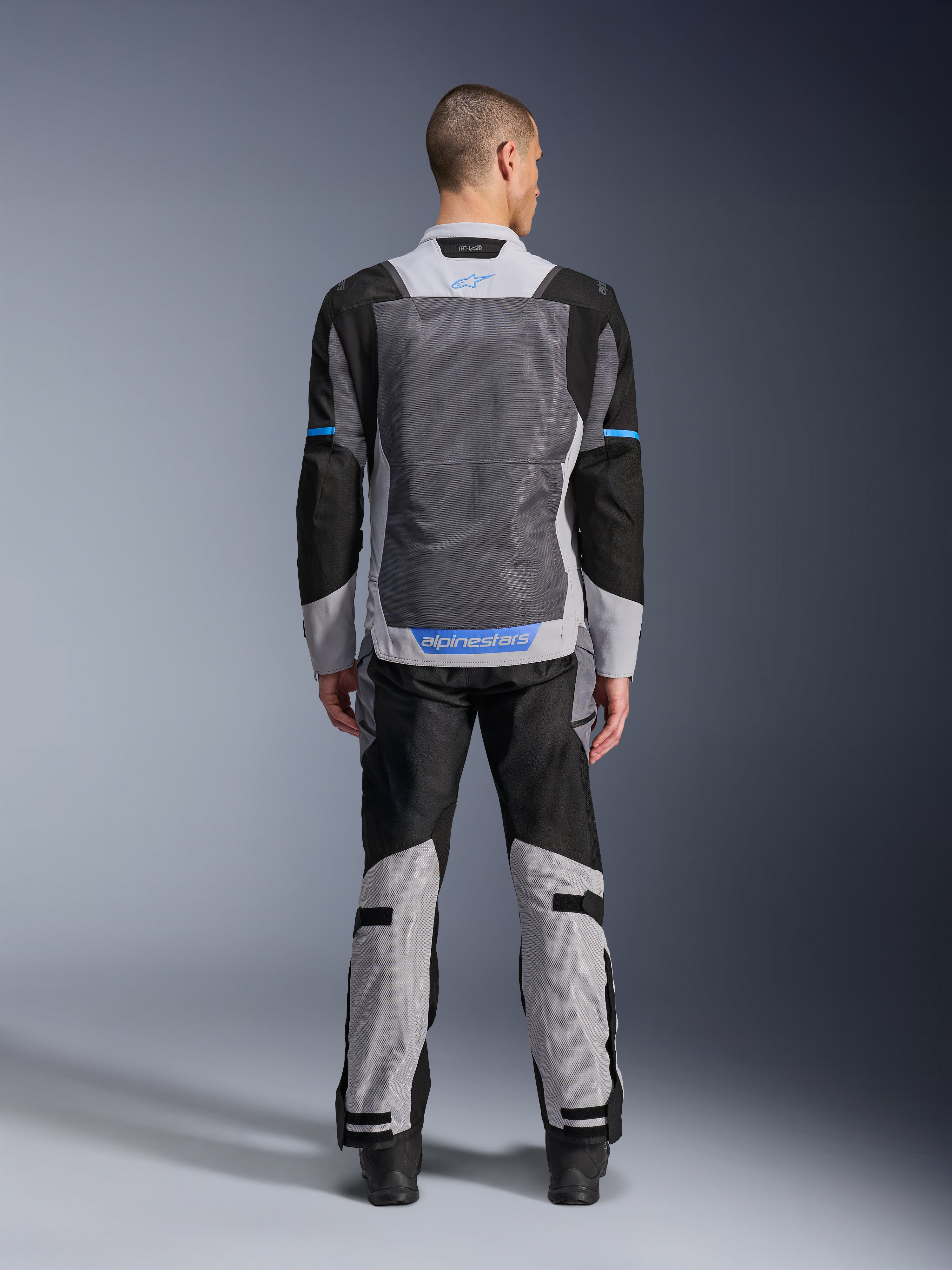 Alpinestars ST-2 Air-jacka, Ice Gray Dark Gray Black, vy bakifrån på modell, med stora ventilationspaneler i mesh, blå detaljer och Alpinestars-logotyp, kombinerad med matchande motorcykelbyxor för adventure touring.