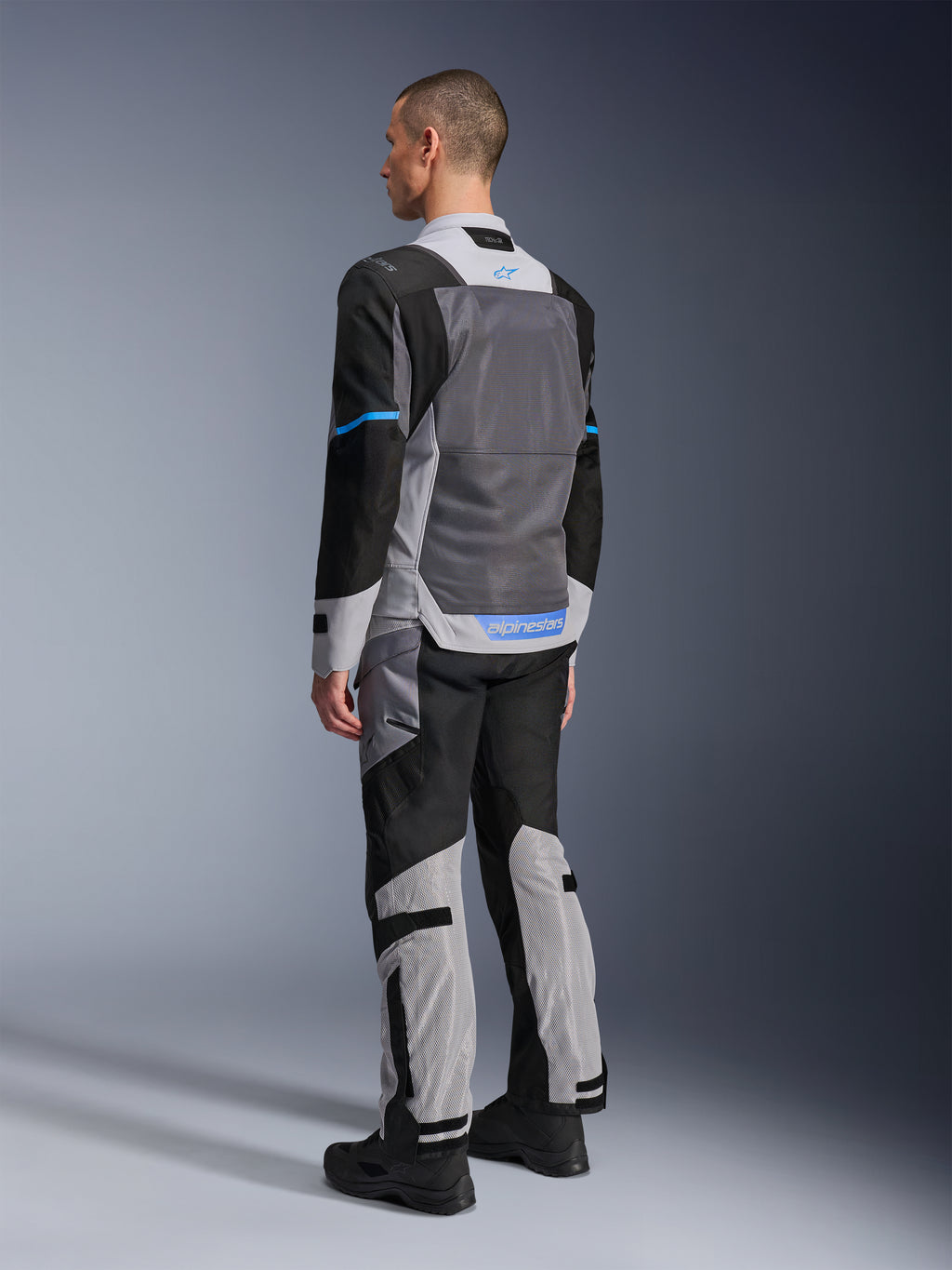 Manlig modell sedd bakifrån bär Alpinestars ST-2 Air Jacket i Ice Gray, Dark Gray och Black, med en stor grå mesh-ryggpanel, blå logotypdetaljer och matchande motorcykelbyxor för sommartouring.