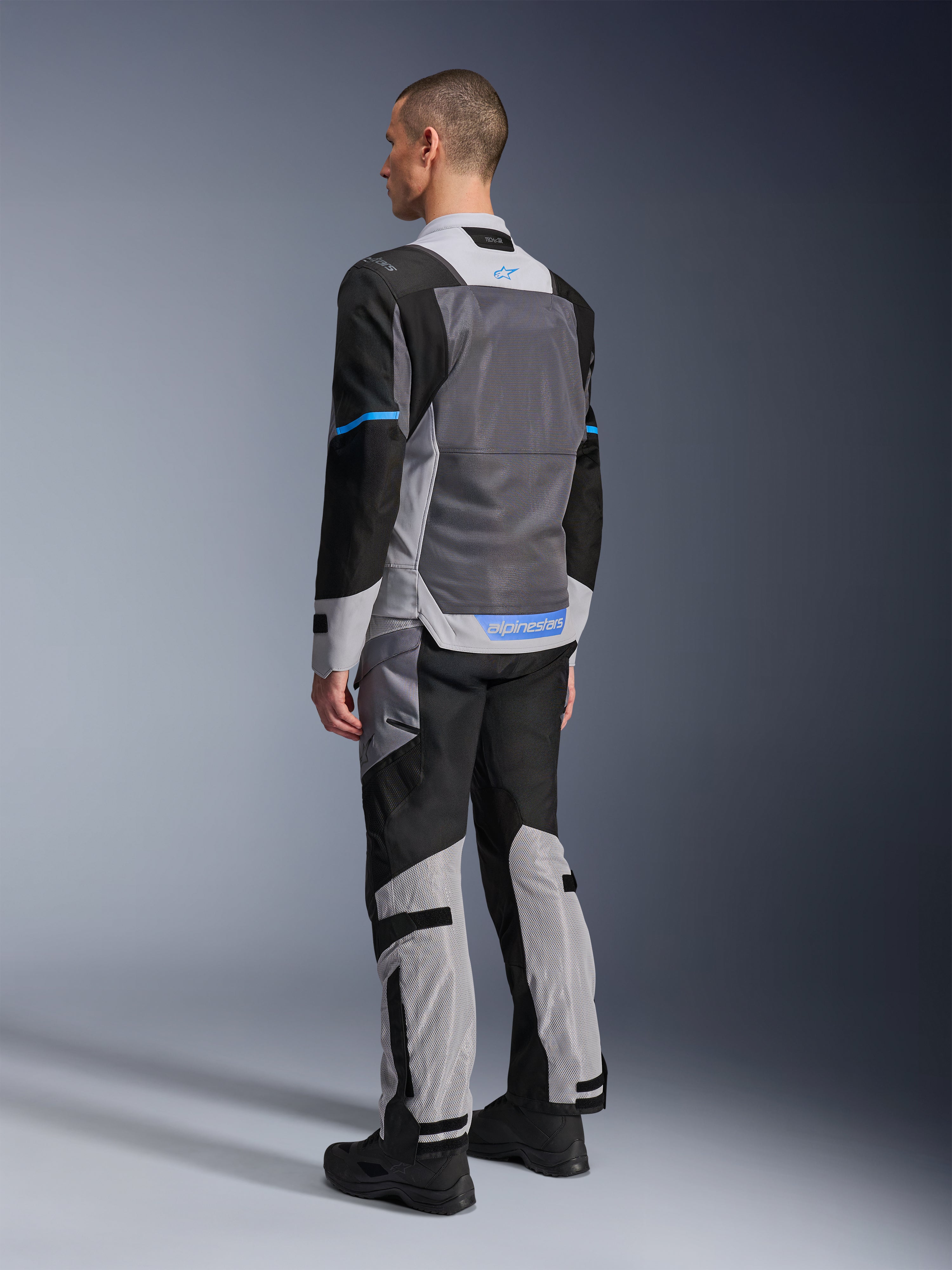 Manlig modell sedd bakifrån bär Alpinestars ST-2 Air Jacket i Ice Gray, Dark Gray och Black, med en stor grå mesh-ryggpanel, blå logotypdetaljer och matchande motorcykelbyxor för sommartouring.