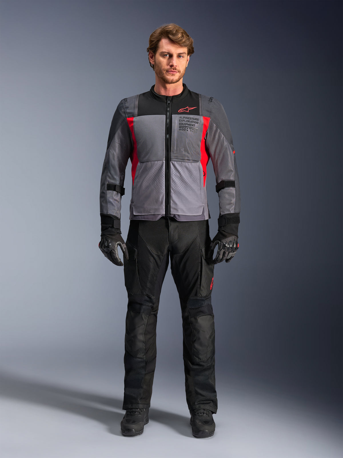 Alpinestars ST-2 Air Jacket in Deep Gray and Bright Red, worn by a male model with black motorcycle pants and gloves, featuring large mesh panels for airflow and modern exploration equipment branding