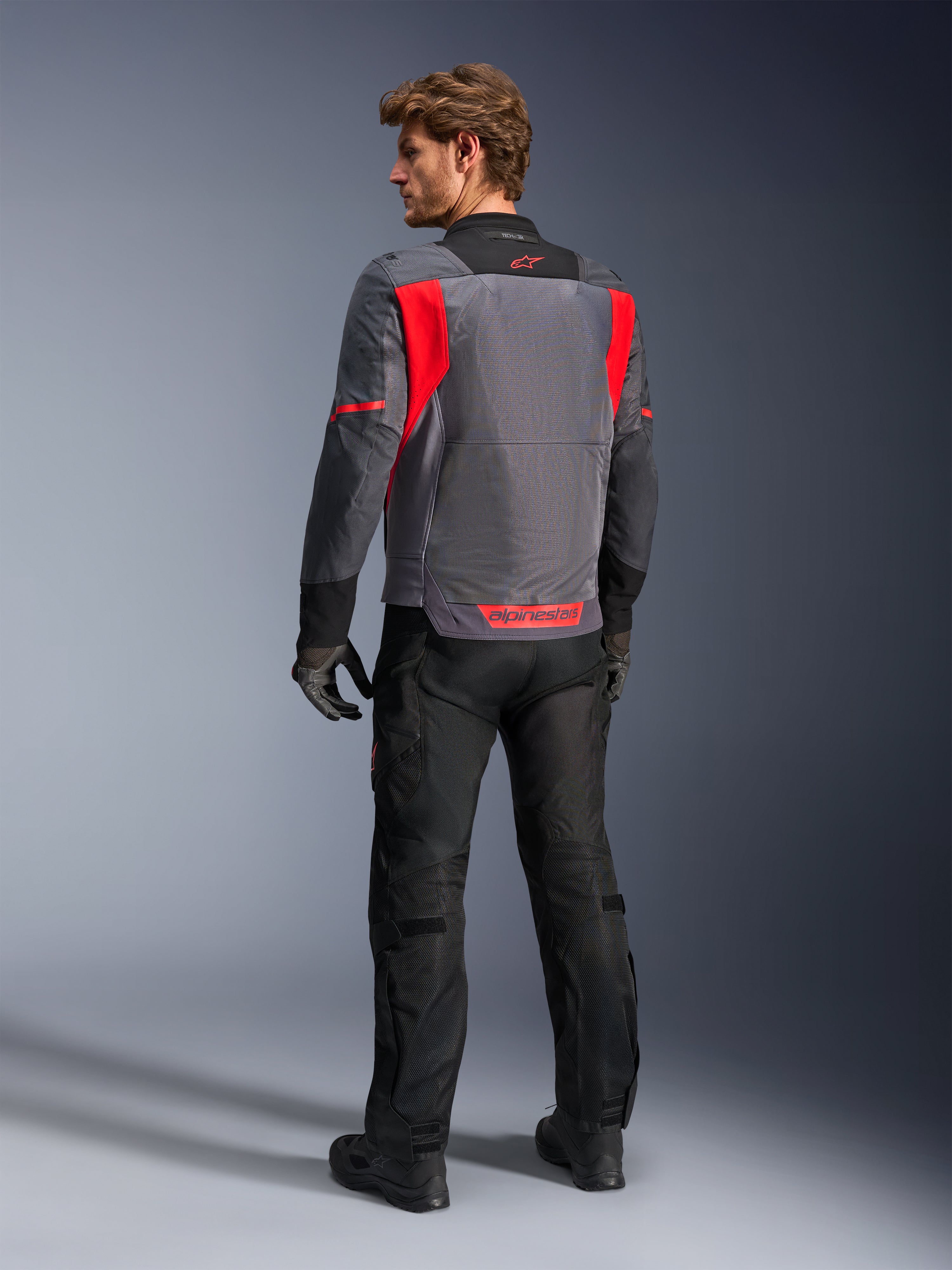 Male rider from behind wearing the Alpinestars St-2 Air Jacket in Deep Gray, Dark Gray and Bright Red, showcasing the large mesh ventilation panel on the back and red branding, styled with black motorcycle pants and gray gloves.