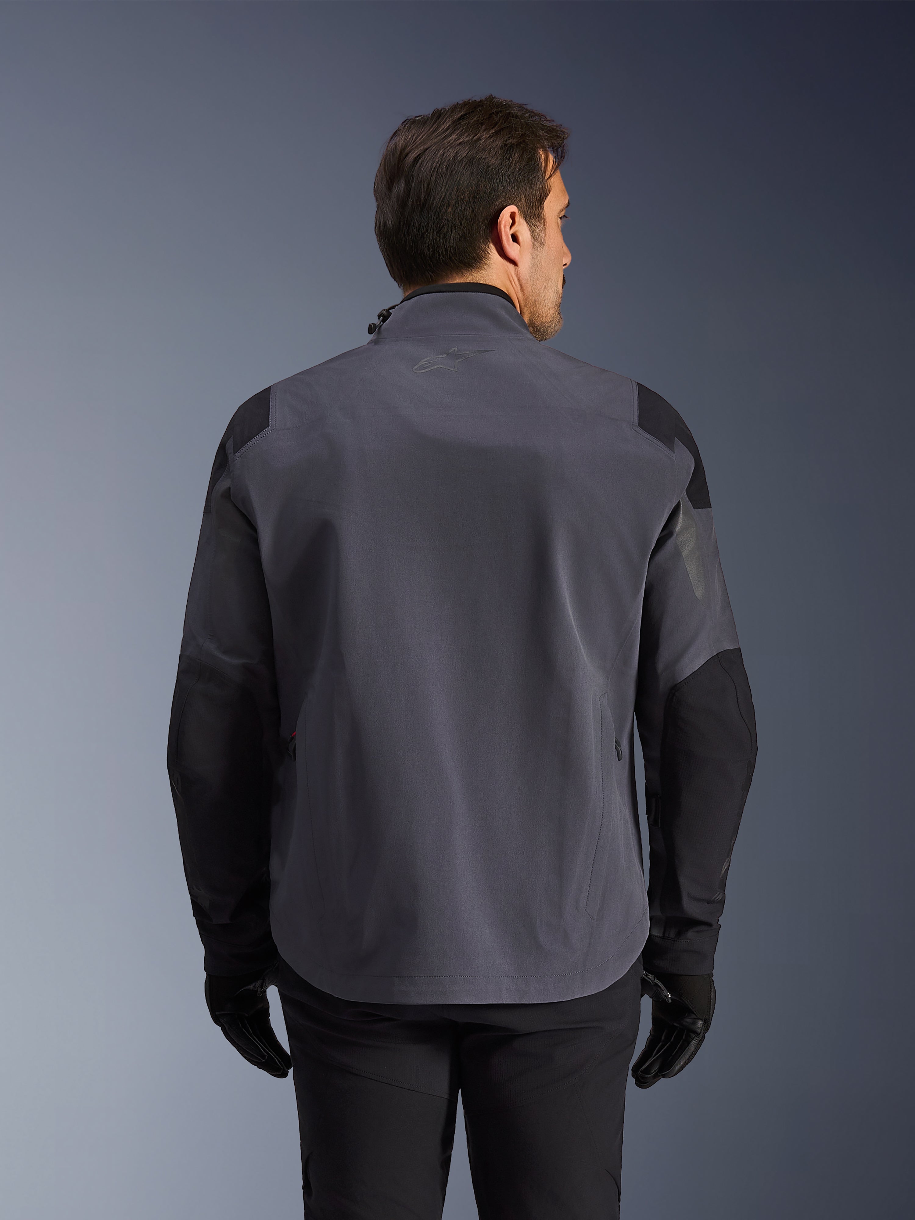Alpinestars Maxdura Drystar®XF Laminated Armored Shell, teknisk äventyrsjacka, mörkgrå och svart, baksida med förstärkta axel- och armbågspaneler, diskret logotyp på övre delen av ryggen, buren med svarta handskar och byxor, konstruerad för skydd i alla väder