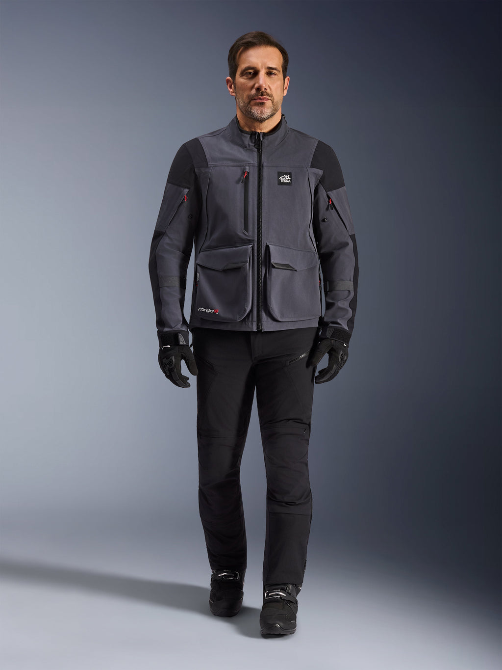 Maxdura Drystar®XF Laminated Armored Shell, motorcykeljacka, Deep Gray Black, bärs av en modell med svarta byxor och handskar, med stora utanpåfickor, röda dragkedjedetaljer och All Terra-branding, teknisk utrustning för äventyrskörning