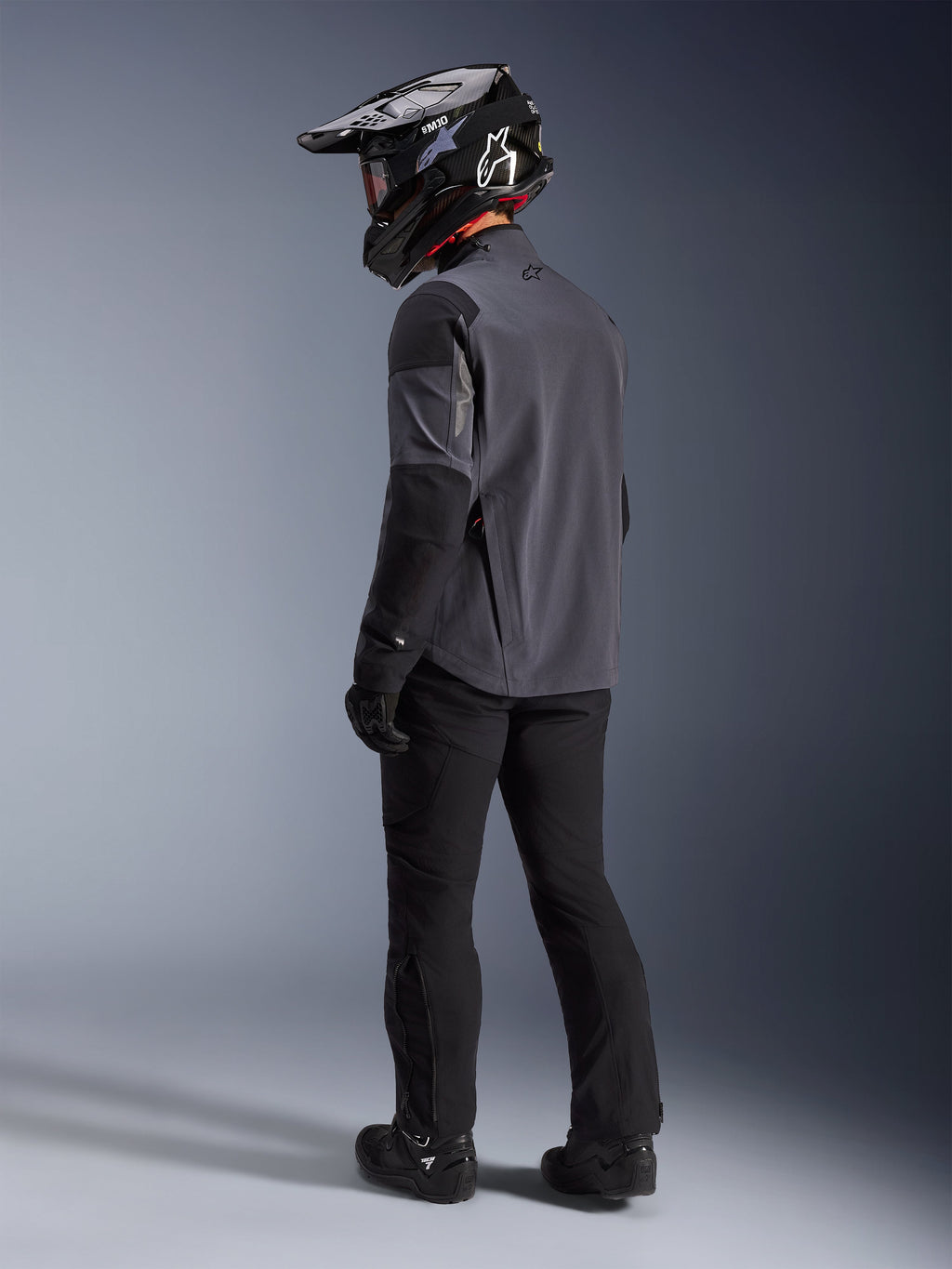 Äventyrsförare iklädd Maxdura Drystar®XF Laminated Armored Shell i Deep Gray Black, ryggvy. Komplett utrustning inklusive svarta motorcykelbyxor, en svart motocrosshjälm med Alpinestars-märkning och svarta off-road-stövlar. Teknisk åkutrustning för alla väder för dual-sport-prestanda.
