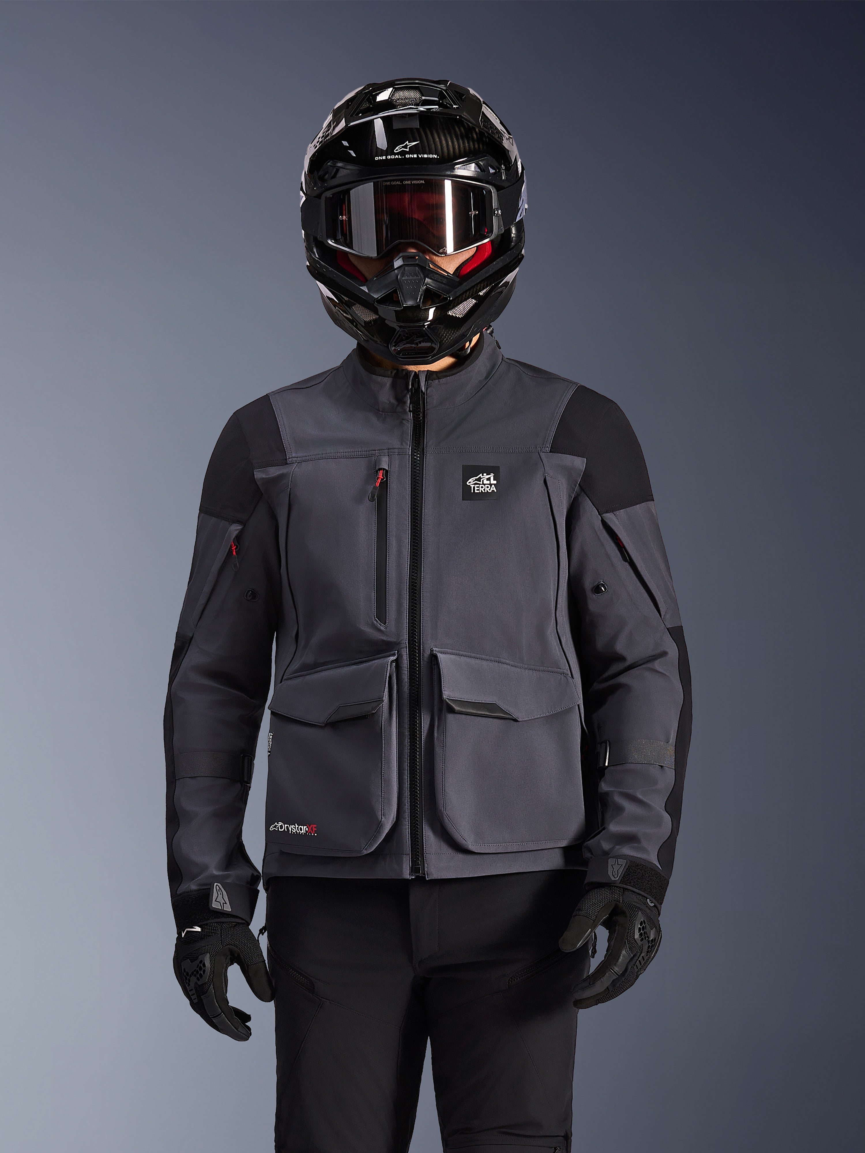 Alpinestars Maxdura Drystar®XF Laminated Armored Shell, motorcykeljacka, mörkgrå och svart, bärs av en förare med svart hjälm, goggles och handskar, med stora tekniska cargofickor och ALL TERRA-märkning, adventure touring-utrustning