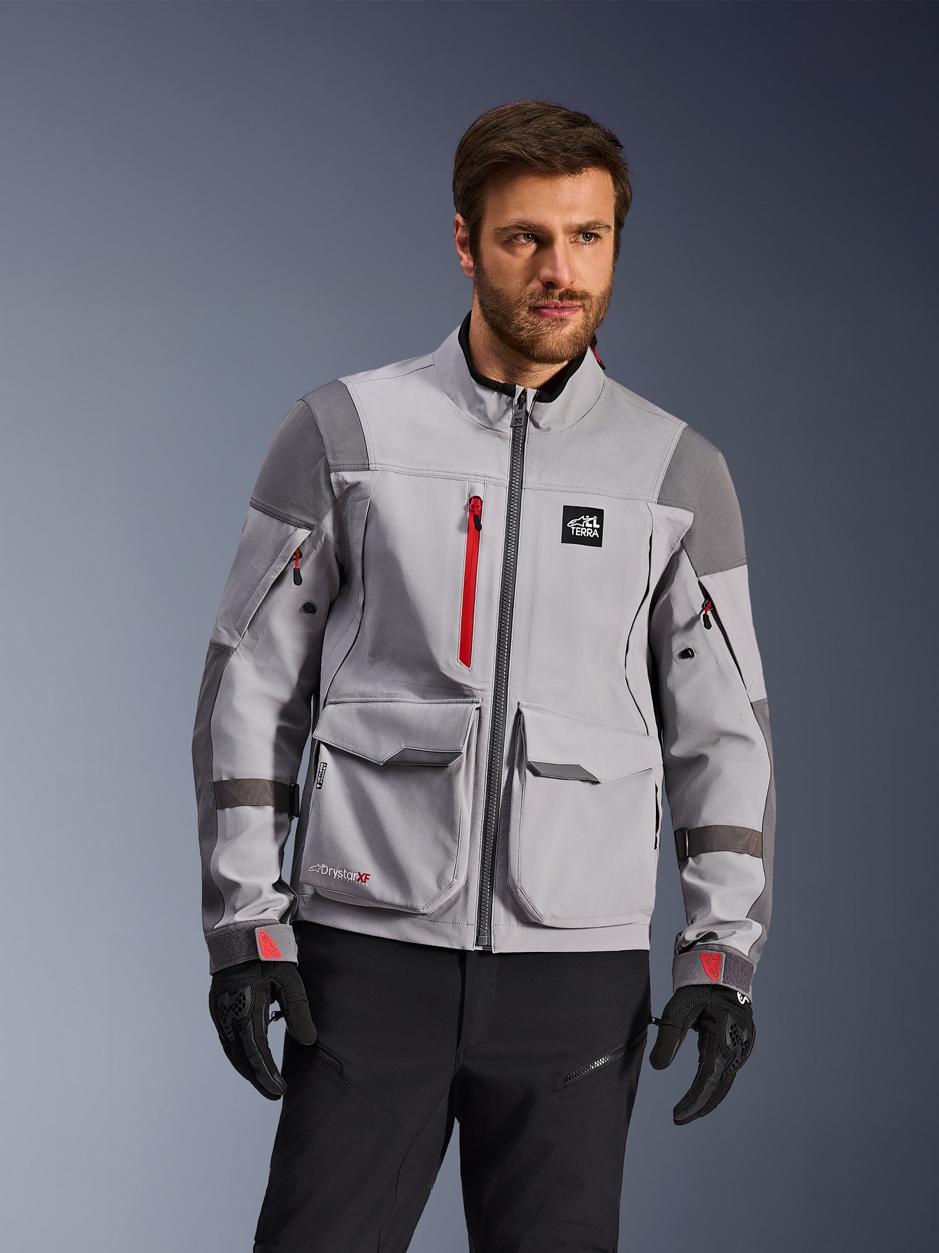 Alpinestars Maxdura Drystar®XF Laminated Armored Shell, äventyrsmotorcykeljacka, mellangrå och mörkgrå med röda detaljer, bärs av en modell med svarta handskar och byxor, stora lastfickor och vattentäta dragkedjor, professionell mc-utrustning