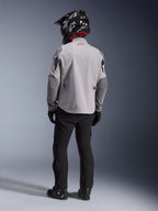 Rear view of a motorcycle rider wearing the Alpinestars Maxdura Drystar®XF Laminated Armored Shell jacket in Mid Gray and Dark Gray, featuring a red star logo on the collar, paired with black adventure pants, a black carbon-patterned helmet, and black boots.