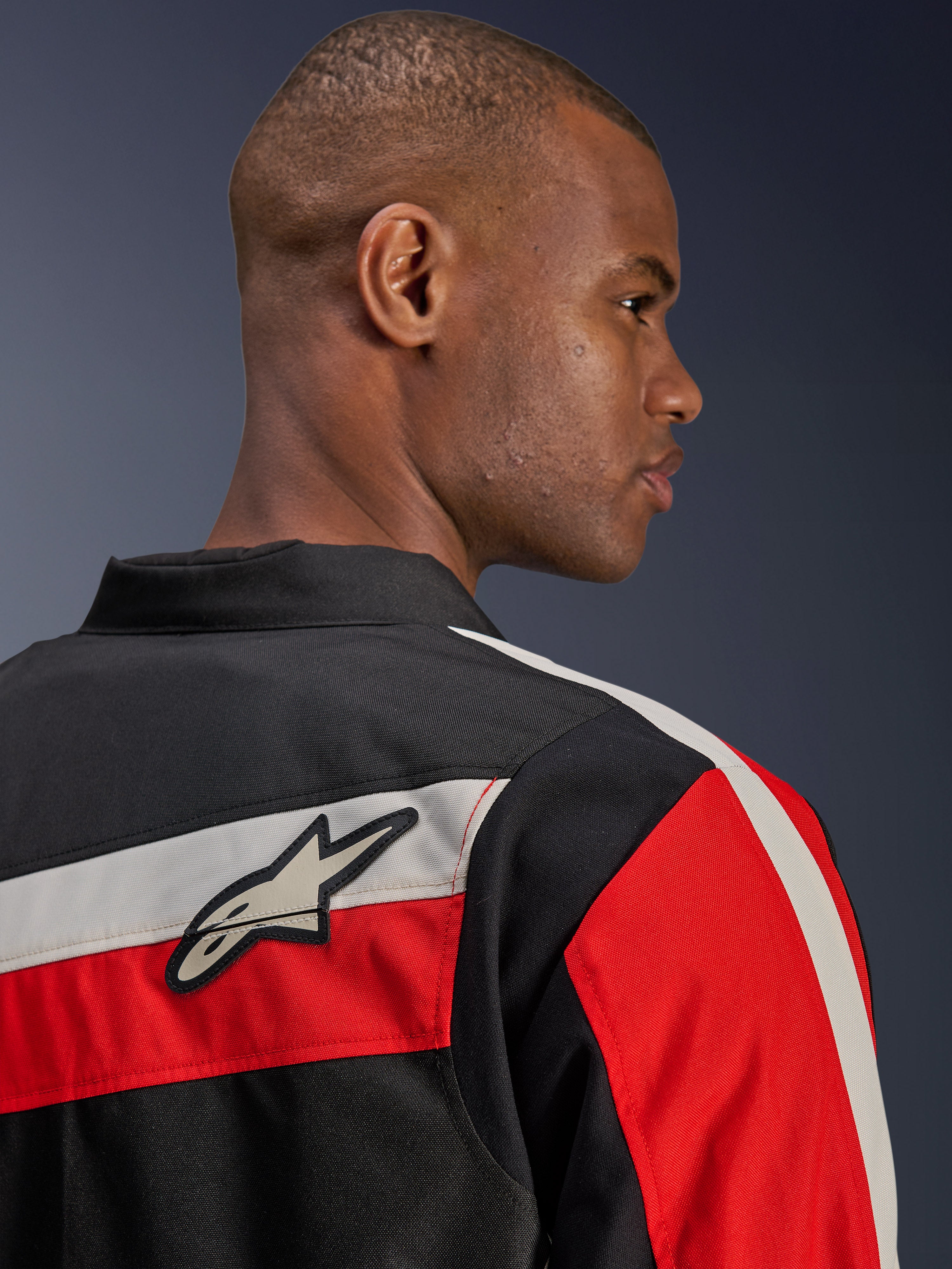 Alpinestars T-Dyno WR-jacka, närbild bakifrån på en modell, textilmotorcykeljacka i svart med horisontella ecru-färgade och klaröda ränder, med Alpinestars stjärnlogotyp på axeln, retroinspirerad bikerstil