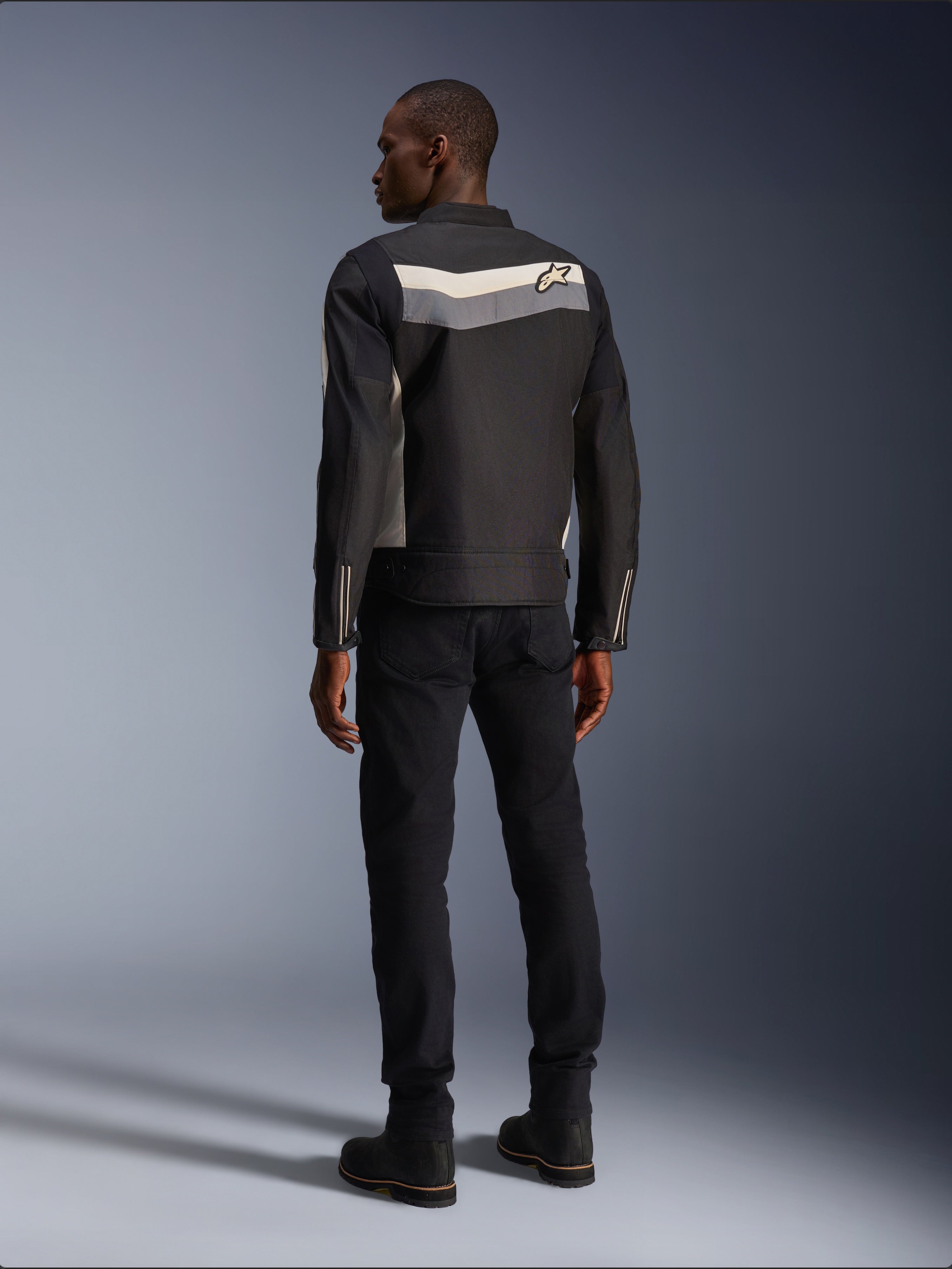 Male model from behind wearing the Alpinestars T-Dyno WR Jacket in Black Dark Gray Ecru, textile motorcycle jacket with a horizontal stripe and star logo on the back, paired with black riding pants and boots, urban motorcycle gear aesthetic.