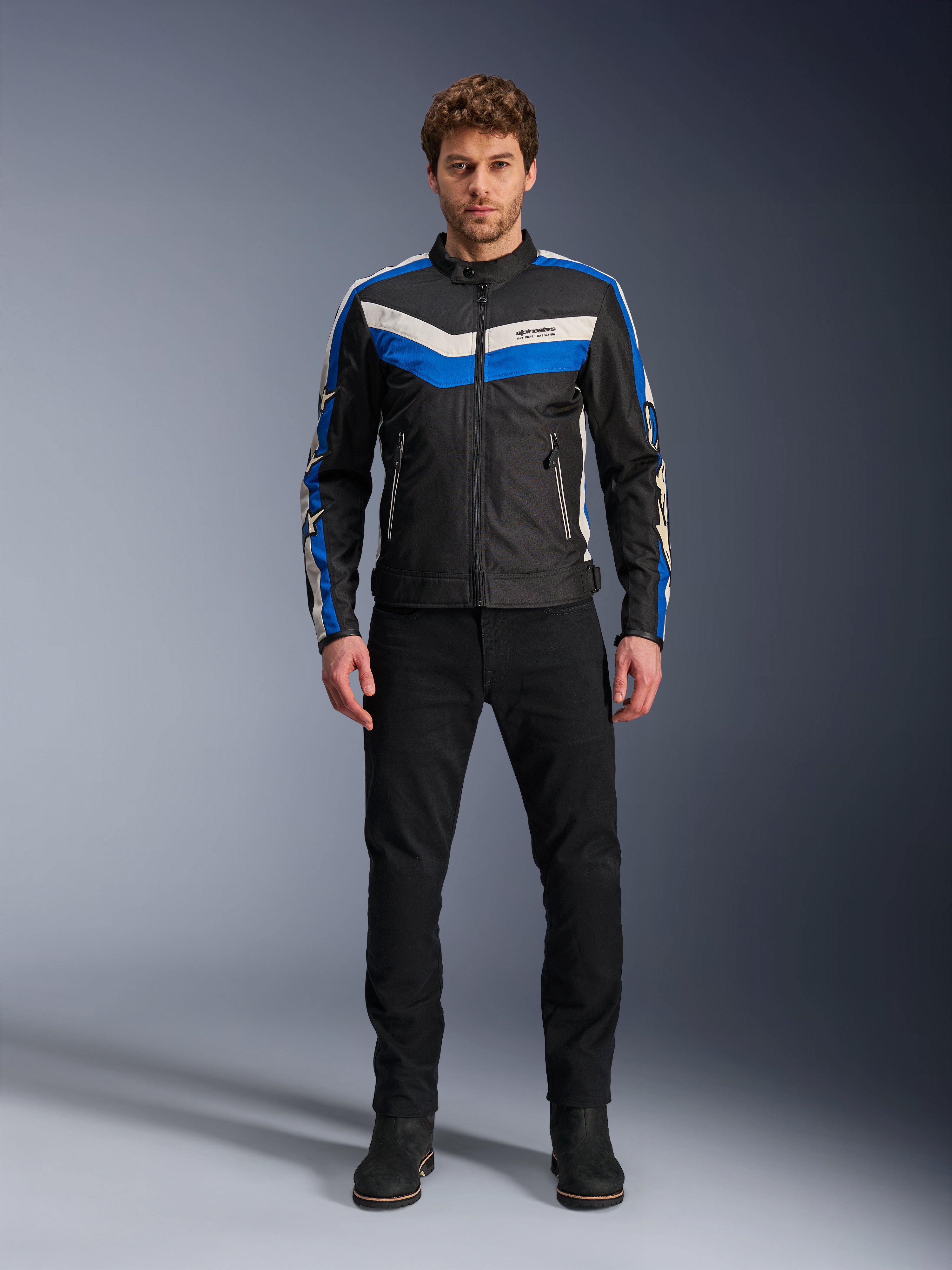 Alpinestars T-Dyno WR-jacka, textiljacka för motorcykel, svart, blå och ecru, bärs av en manlig modell med svarta byxor och stövlar, med chevron-ränder på bröstet och klassisk bikerdesign