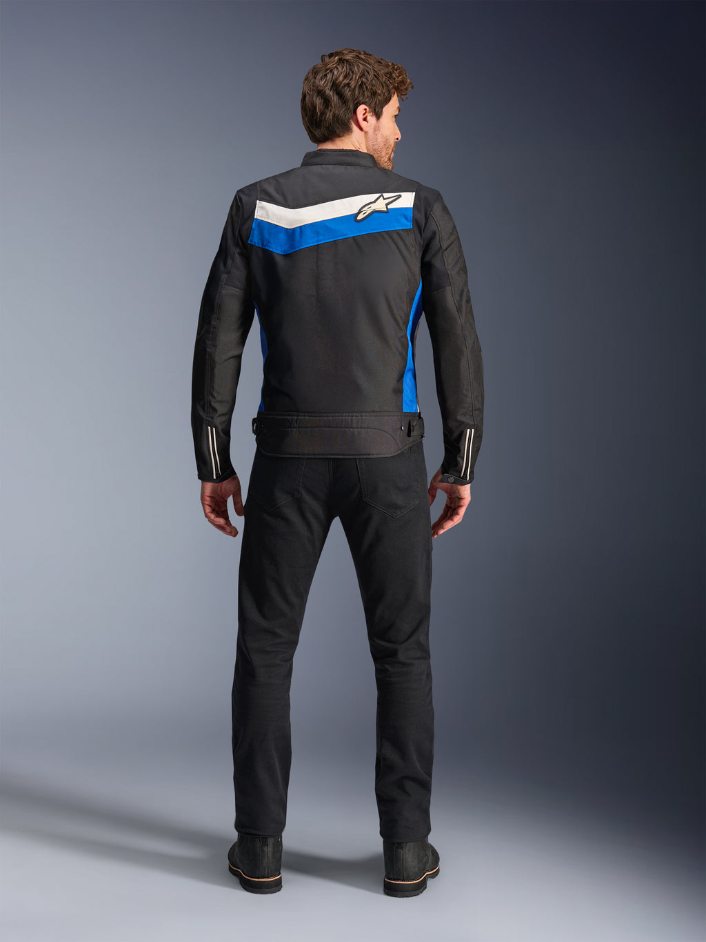 Alpinestars T-Dyno WR Jacket, motorcykeljacka i textil, färgkombinationen Black Blue Ecru, hel ryggvy på modell, med ett blått och off-white chevron-format band med Alpinestars-logotyp, kombinerad med svarta byxor och stövlar, vintageinspirerad urban mc-utrustning